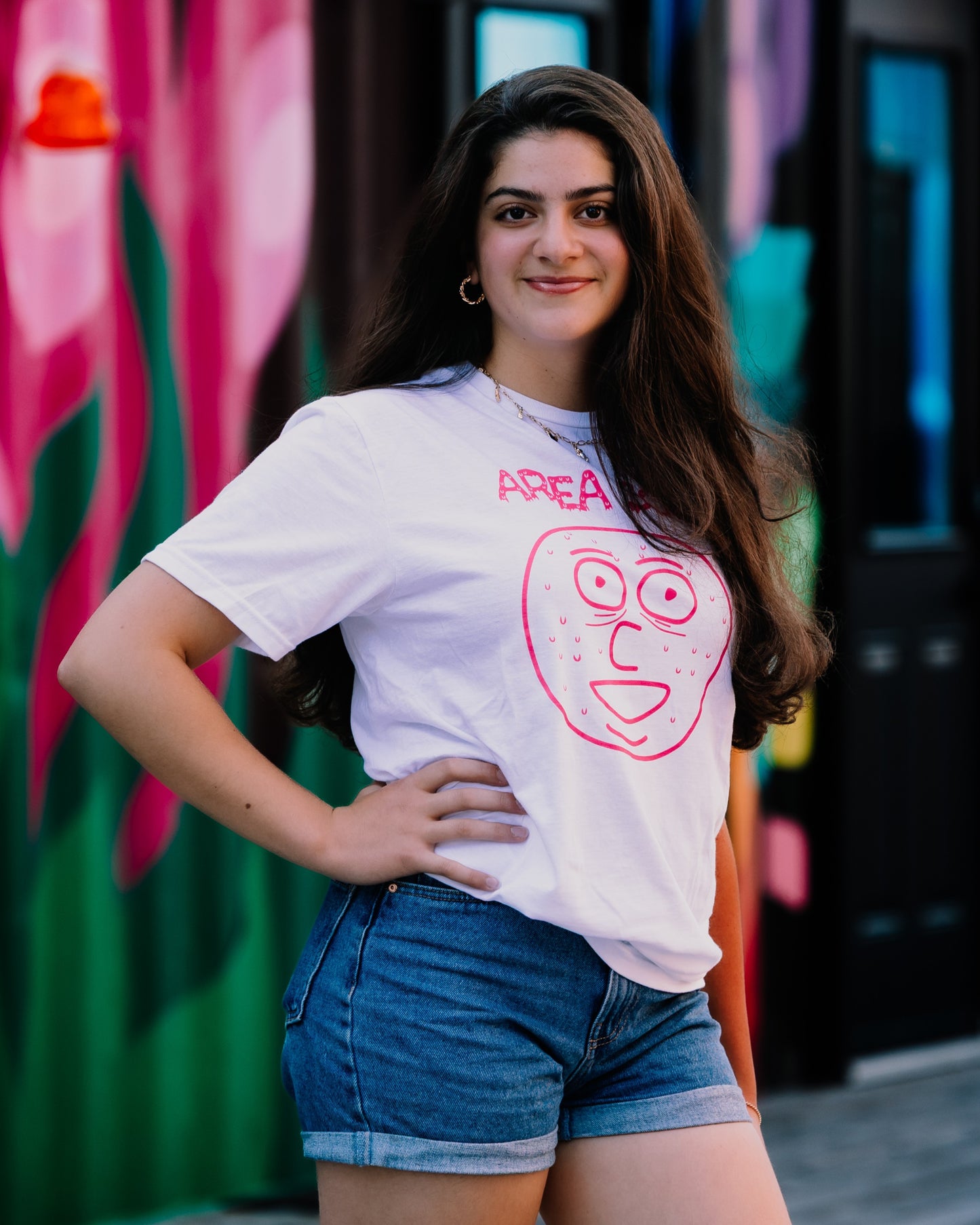 Woman wearing a white t-shirt with a pink graphic design in front of a colourful mural.
