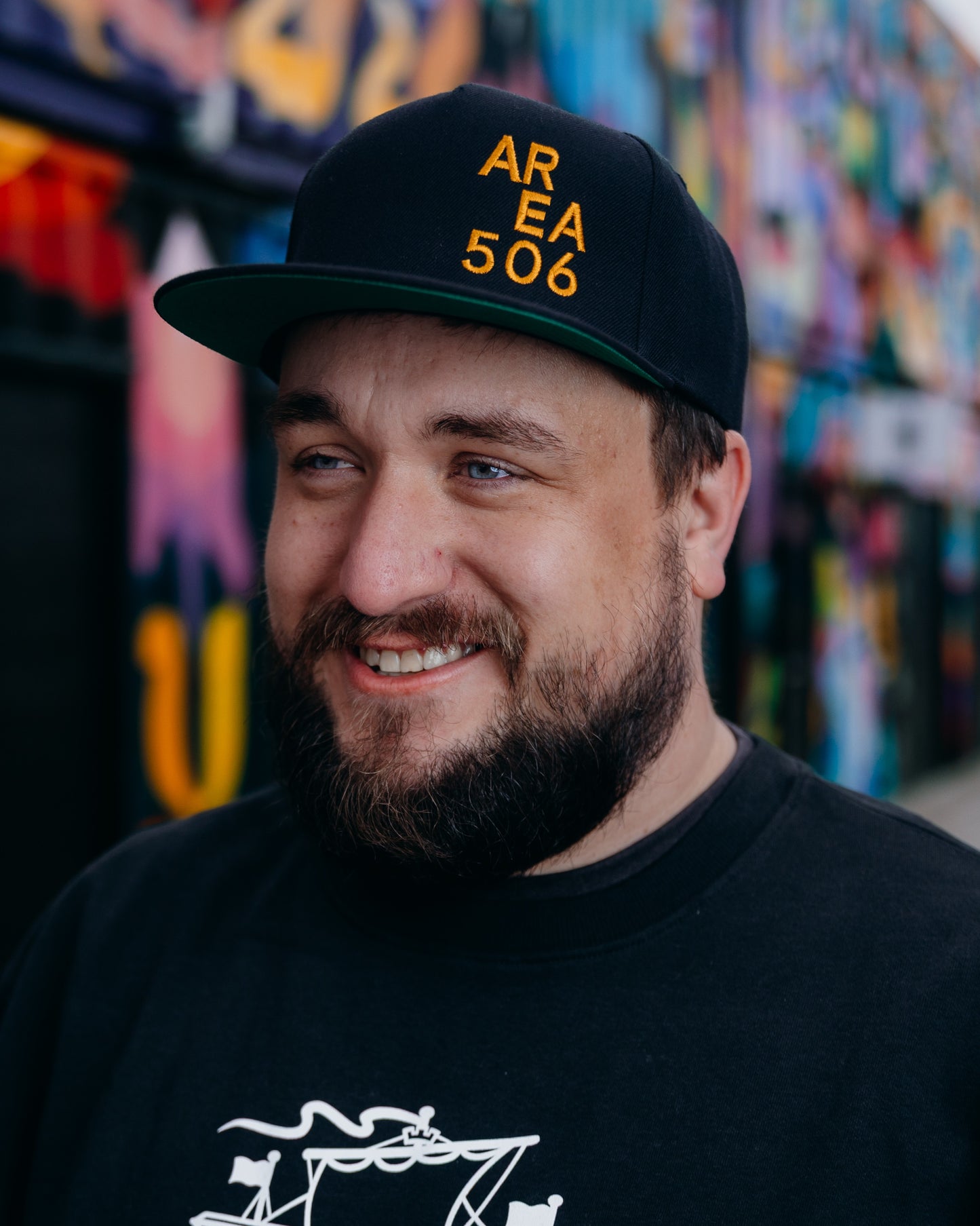 Man wearing a black cap with 'AREA 506' text, smiling in front of a colourful mural.