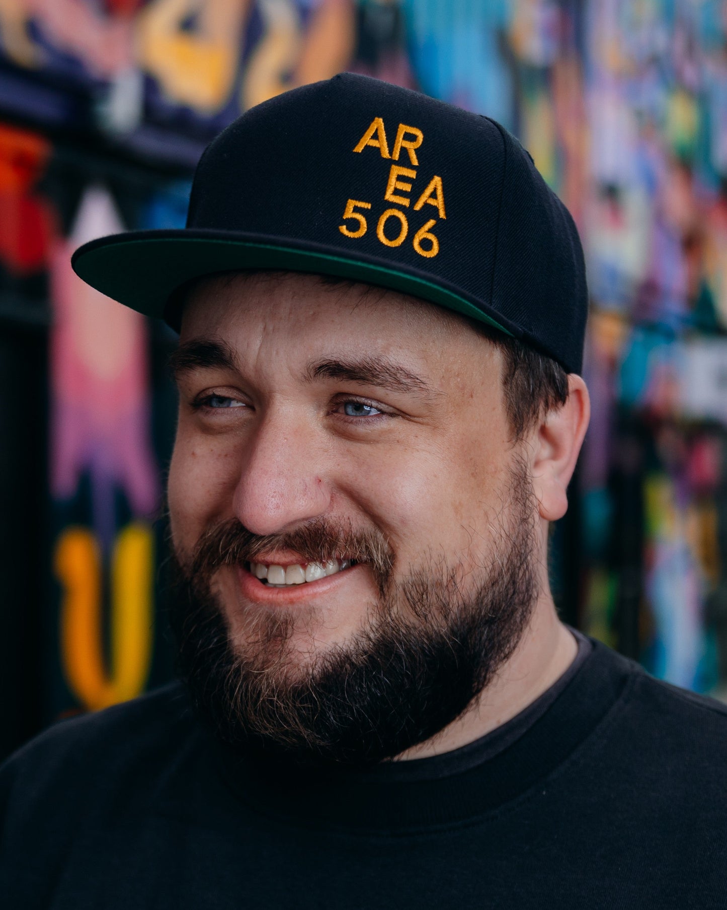 Man wearing a black cap with 'AREA 506' text, smiling in front of a colourful mural.
