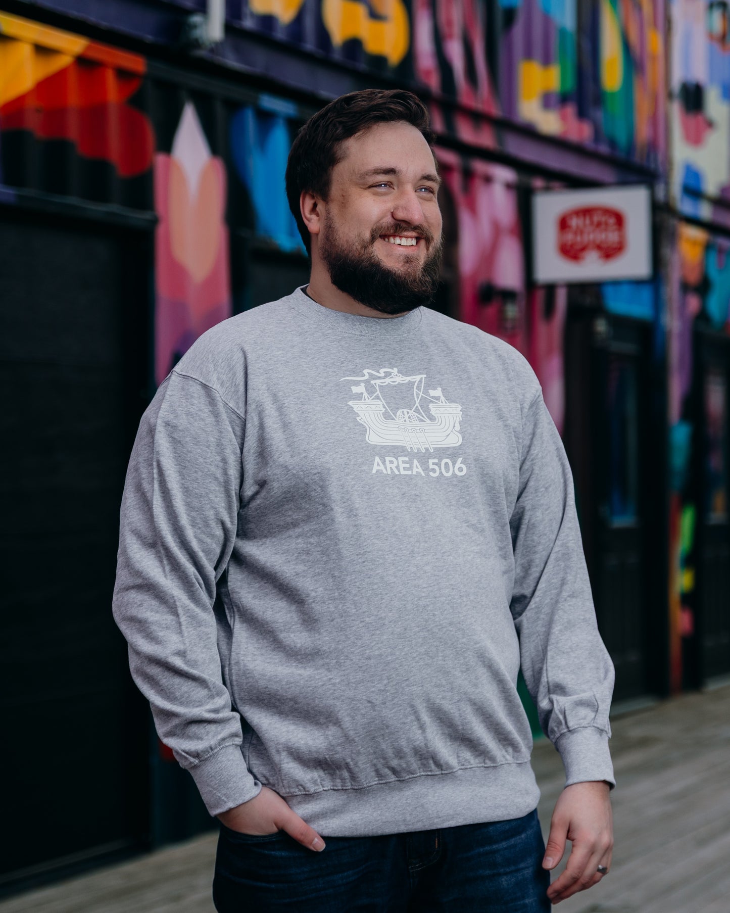 Man wearing a gray sweatshirt with 'AREA 506' print, standing in front of a colourful mural.