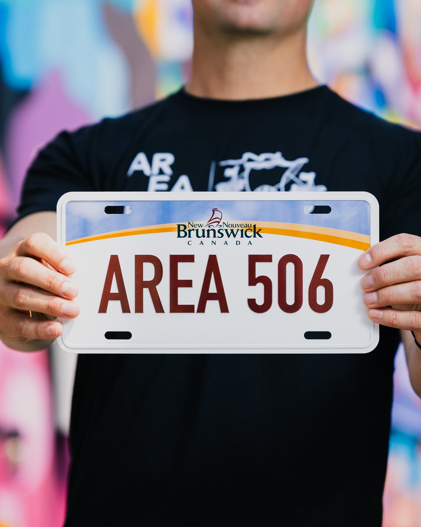 Person holding a tin sign shaped like a license plate with 'AREA 506' and New Brunswick branding.