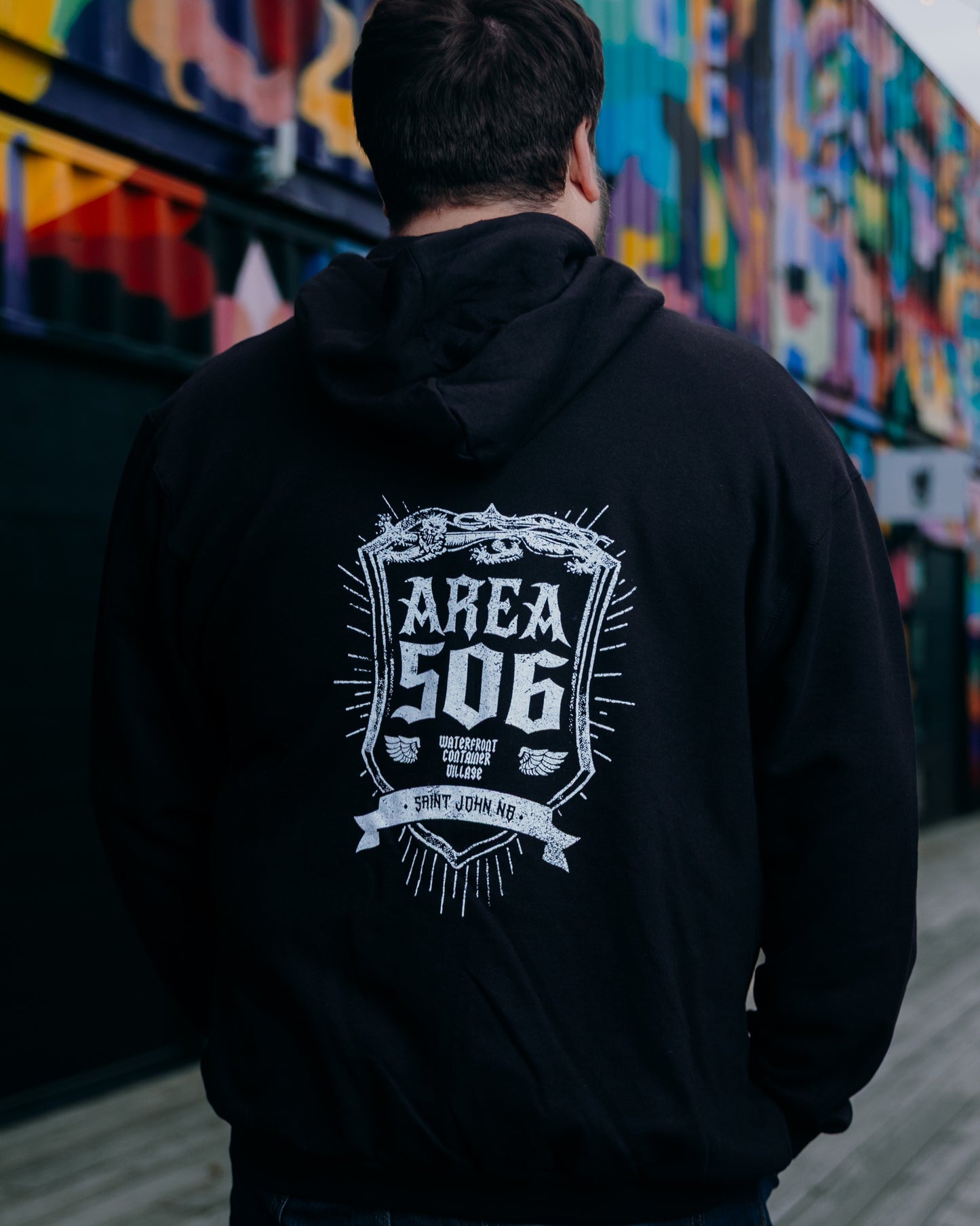 Person wearing a black hoodie with 'AREA 506' logo on the back, standing in front of a colourful mural.