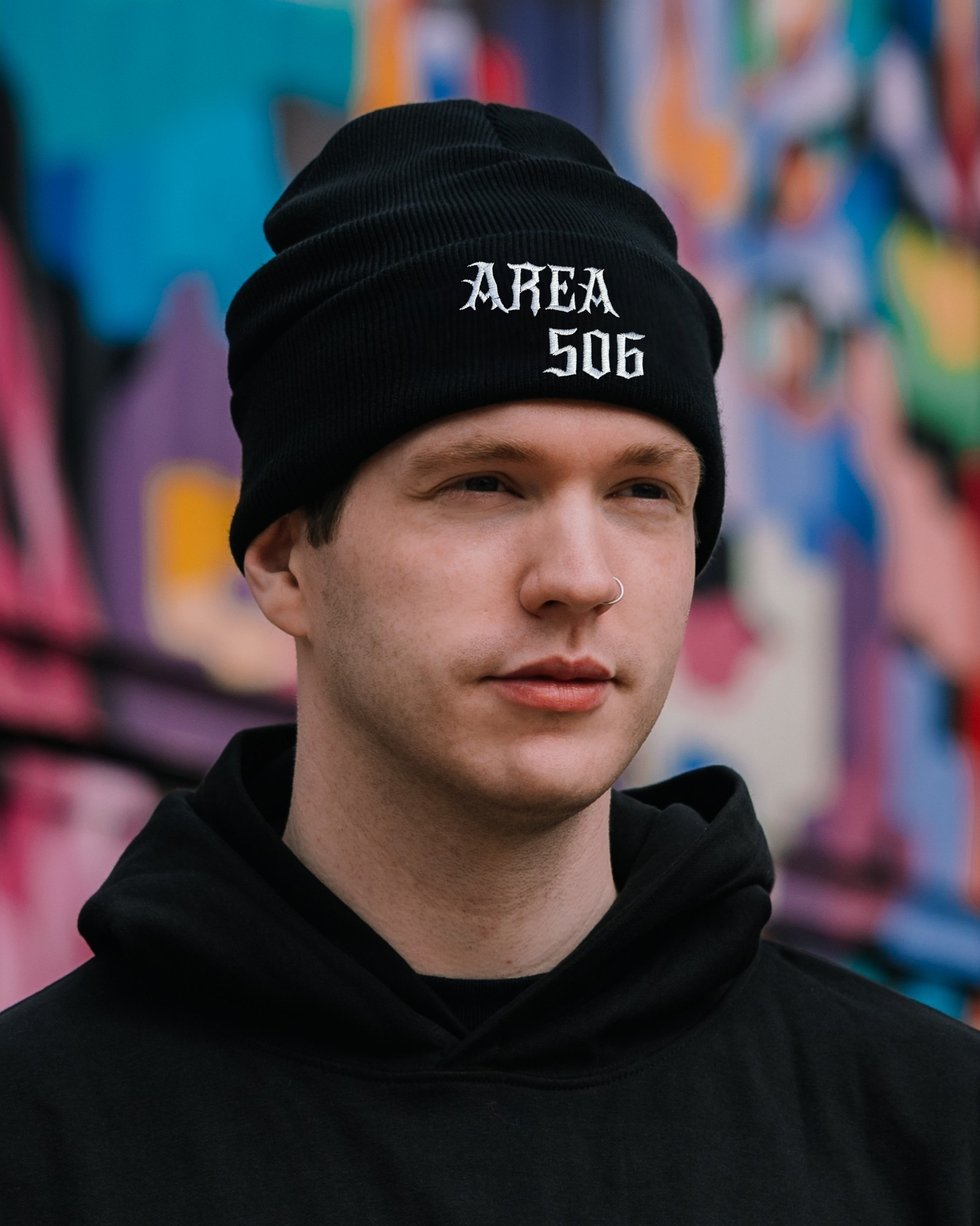 Person wearing a black beanie with 'AREA 506' text in front of a colourful graffiti wall.