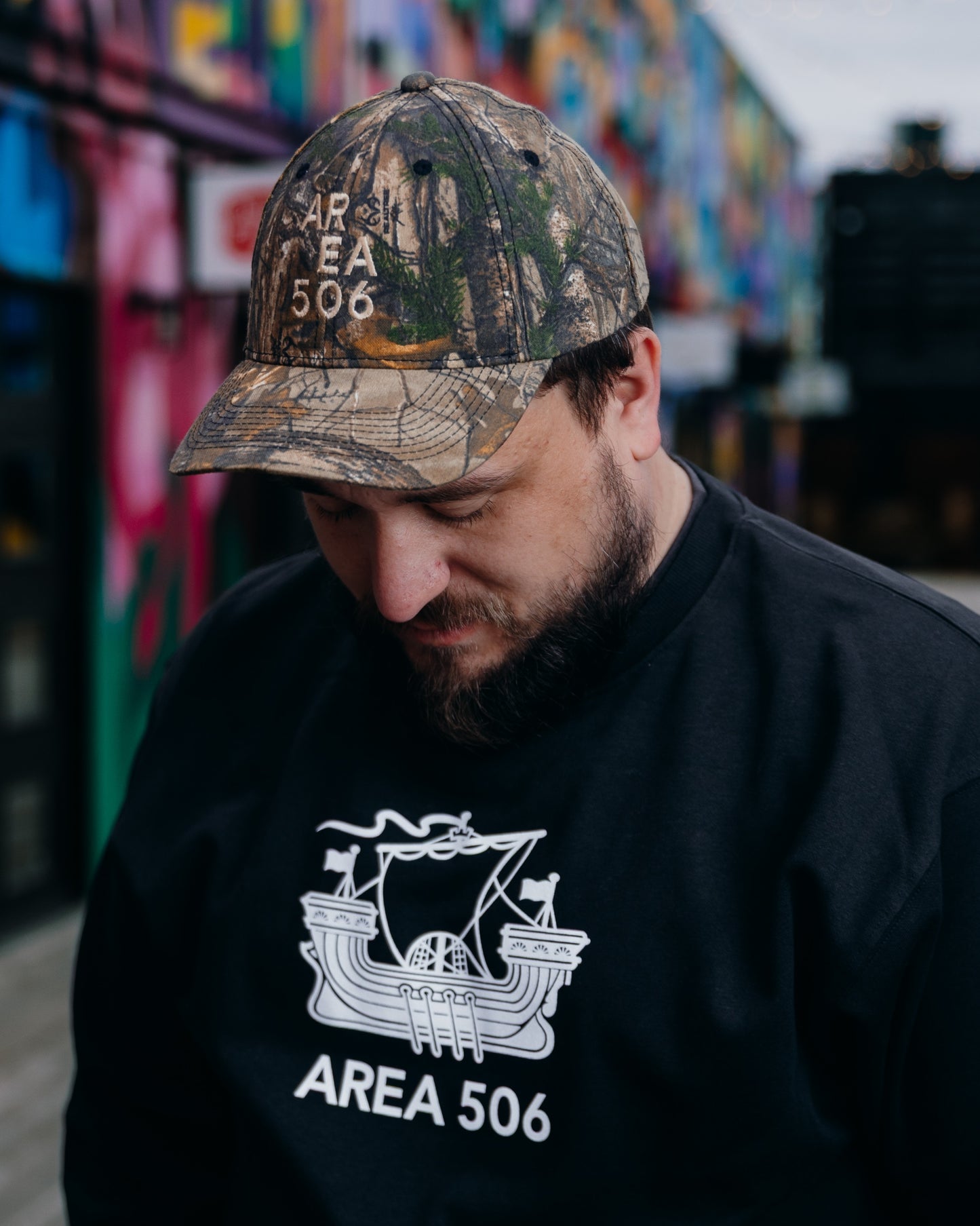 Person wearing a camouflage 'AREA 506' cap and black sweatshirt with logo