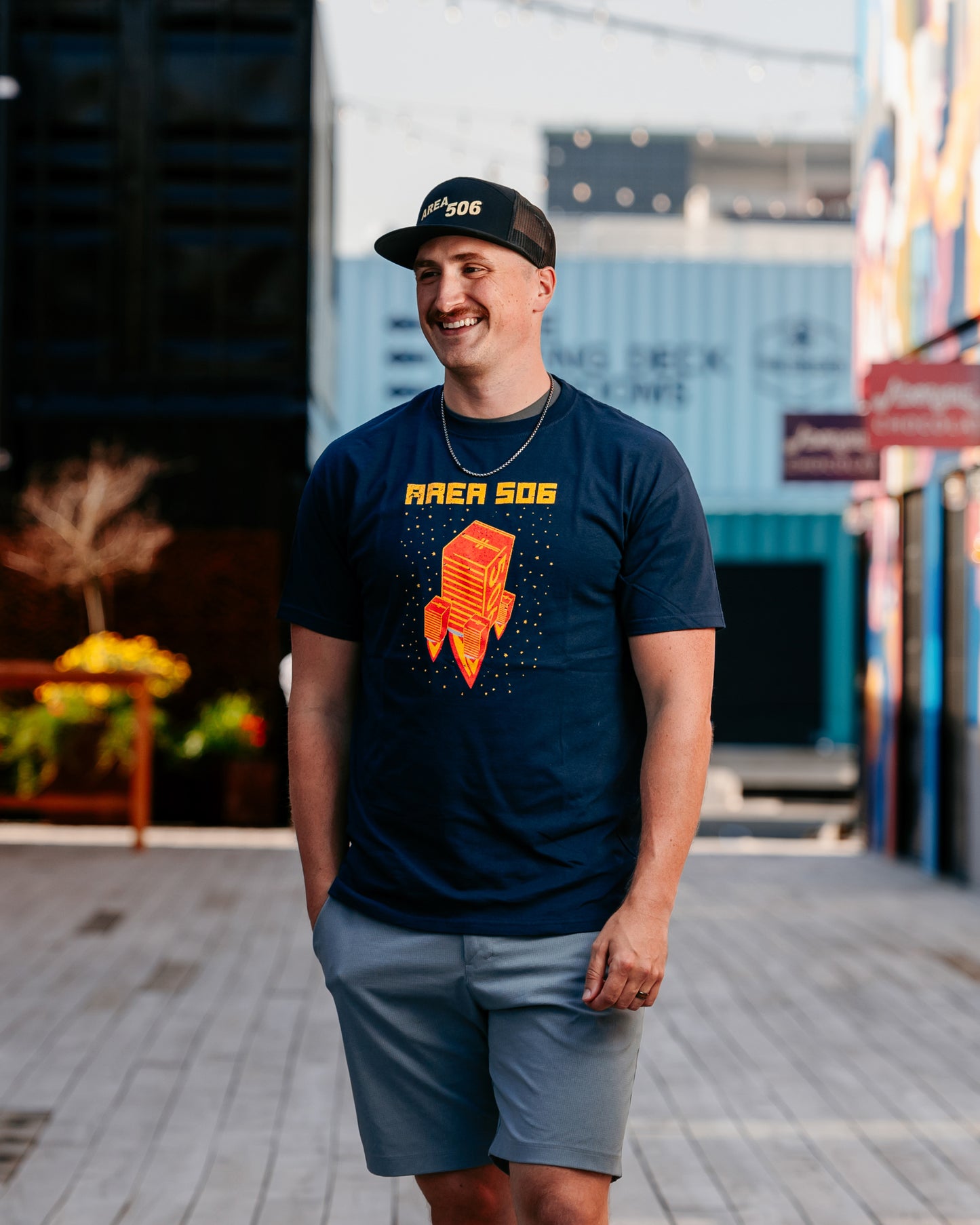 A person wearing a navy blue t-shirt with a red and yellow rocket ship logo, paired with light gray shorts and a black cap, standing on a street with buildings in the background.