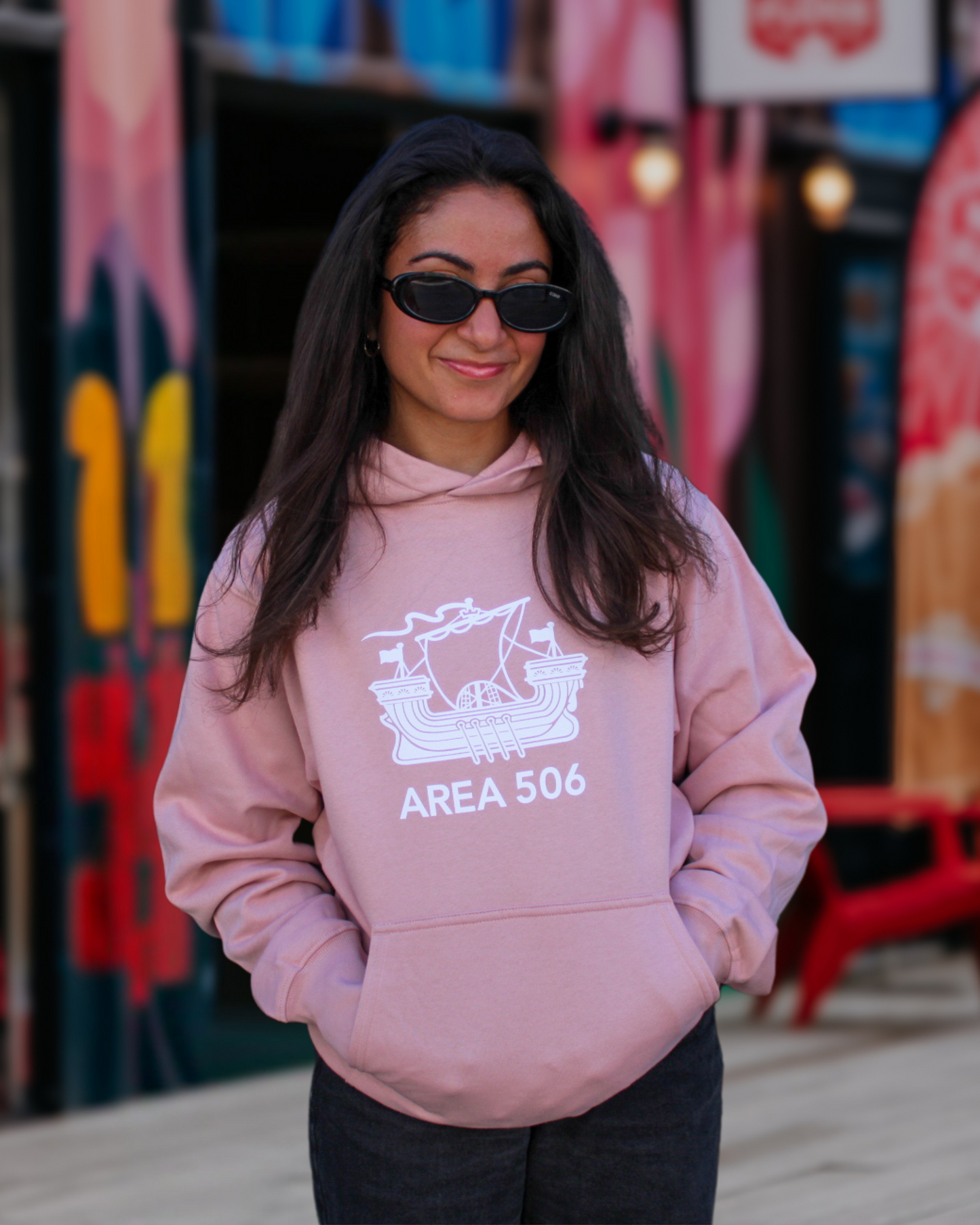 Person wearing a pink hoodie with 'AREA 506' design in front of colourful wall art.