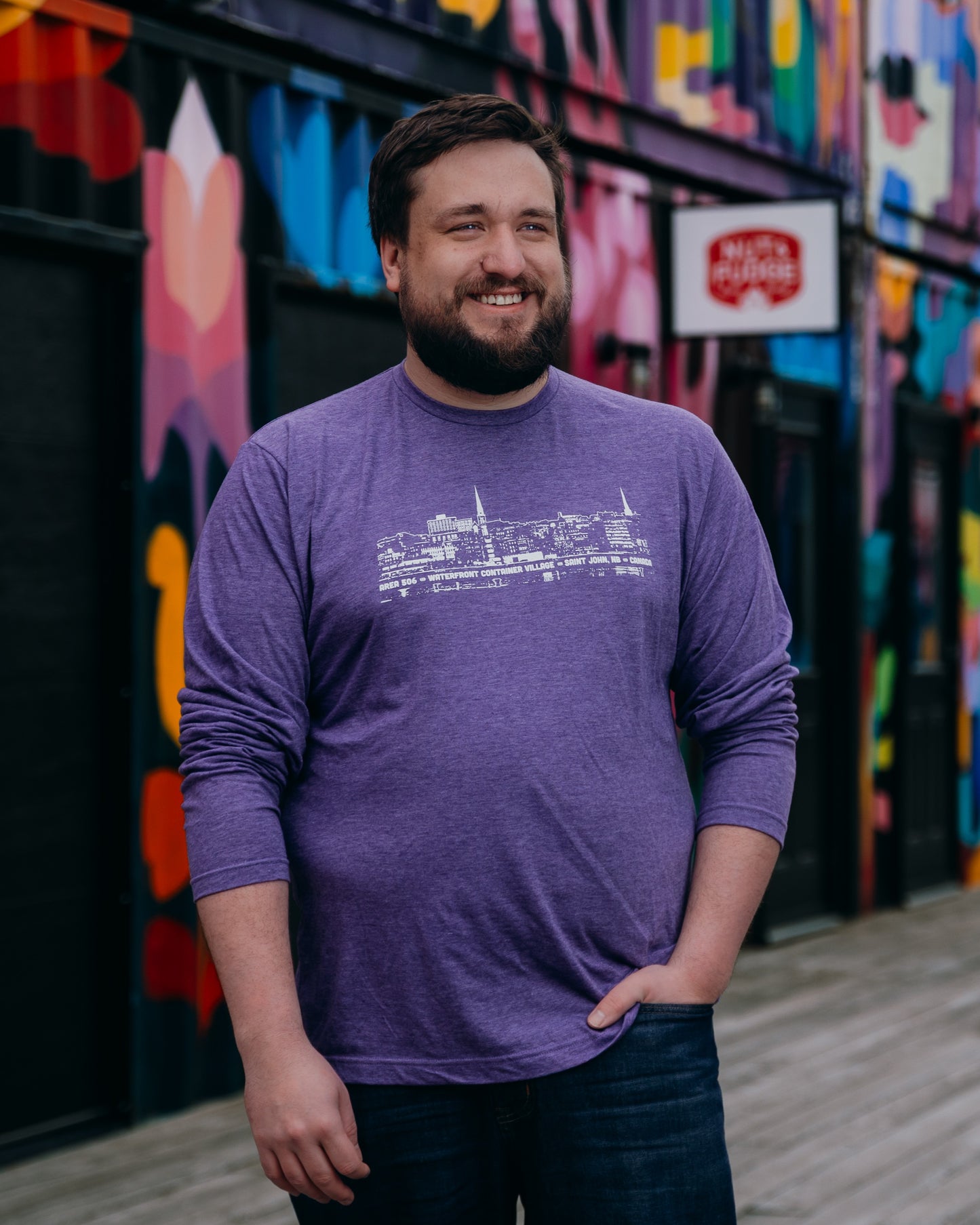 Man wearing a purple long-sleeve shirt with a white graphic design, standing in front of a colourful mural.