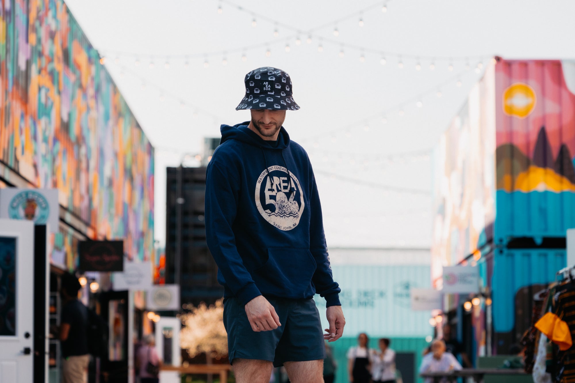 Navy blue pull over hoodie available in three text colours. Green, peach, or blue AREA 506 Waterfront Container Village logo on the front and left sleeve.