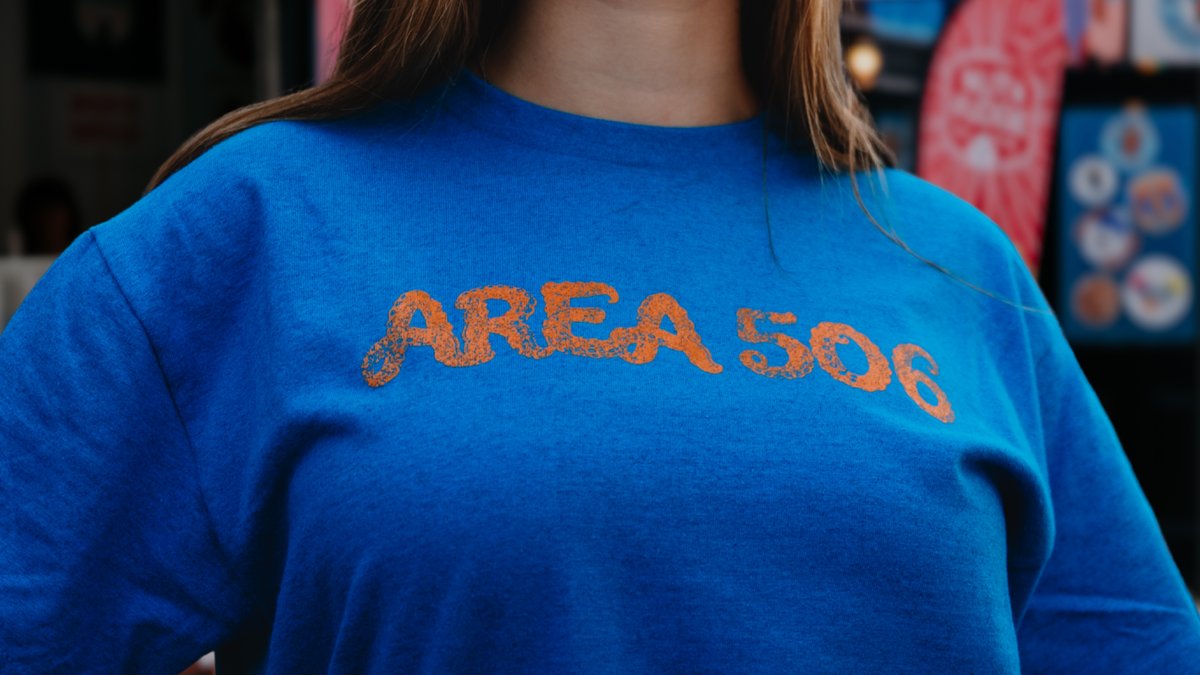 A close-up of a person wearing a bright blue t-shirt with 'AREA 506' printed across the chest in a textured orange font.