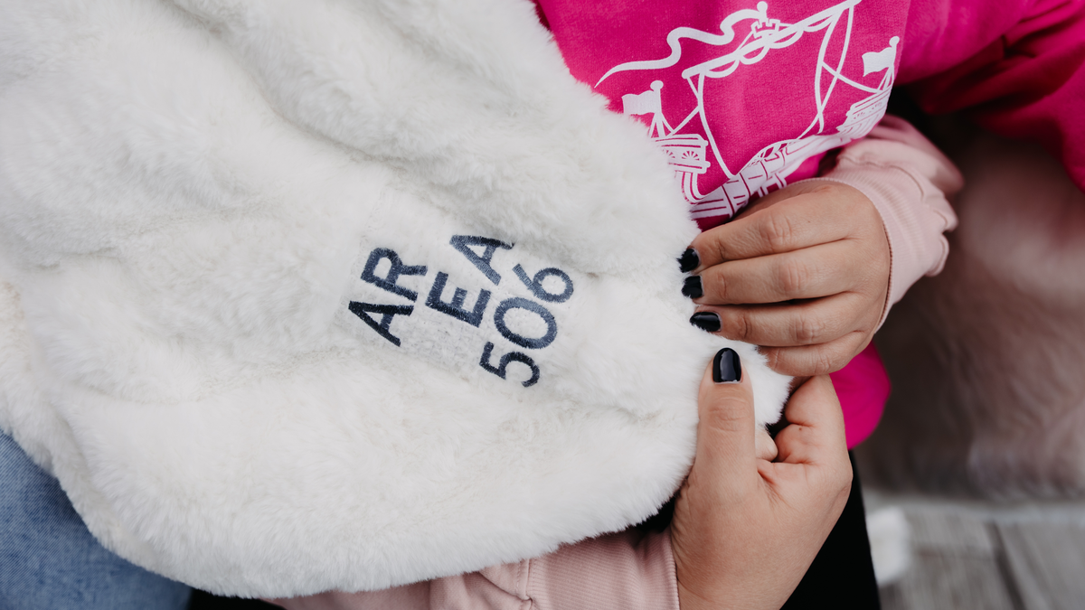 A close-up of a white faux fur blanket or throw with 'AREA 506' embroidered in navy blue, held by a person wearing a pink sweatshirt.