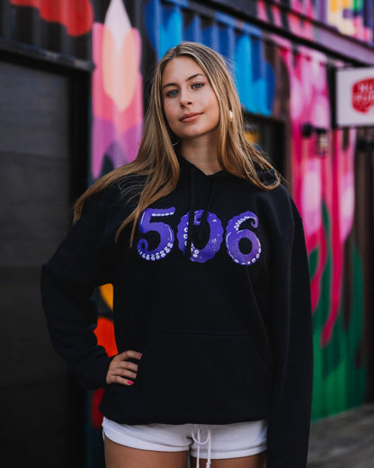 A woman wearing a black hoodie with a purple '506' graphic on the front, standing in front of a colourful graffiti wall.
