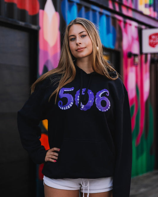 A woman wearing a black hoodie with a purple '506' graphic on the front, standing in front of a colourful graffiti wall.