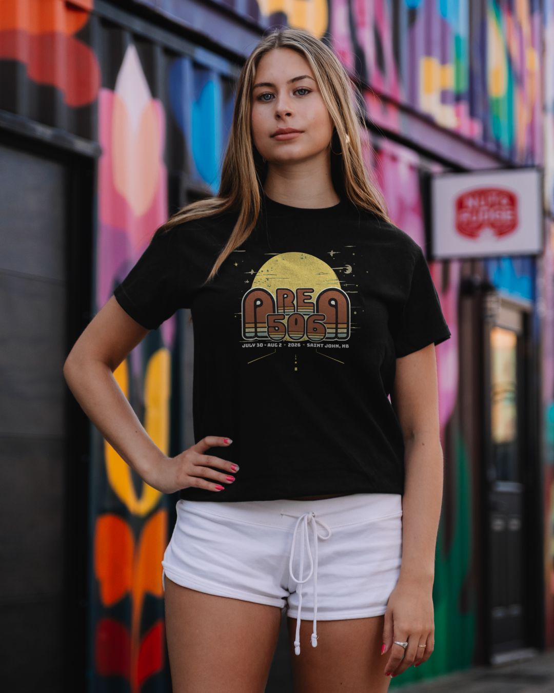 A woman modeling a black graphic t-shirt with a retro-style 'AREA 506' sunset design, paired with white shorts, standing in front of a colourful graffiti wall.