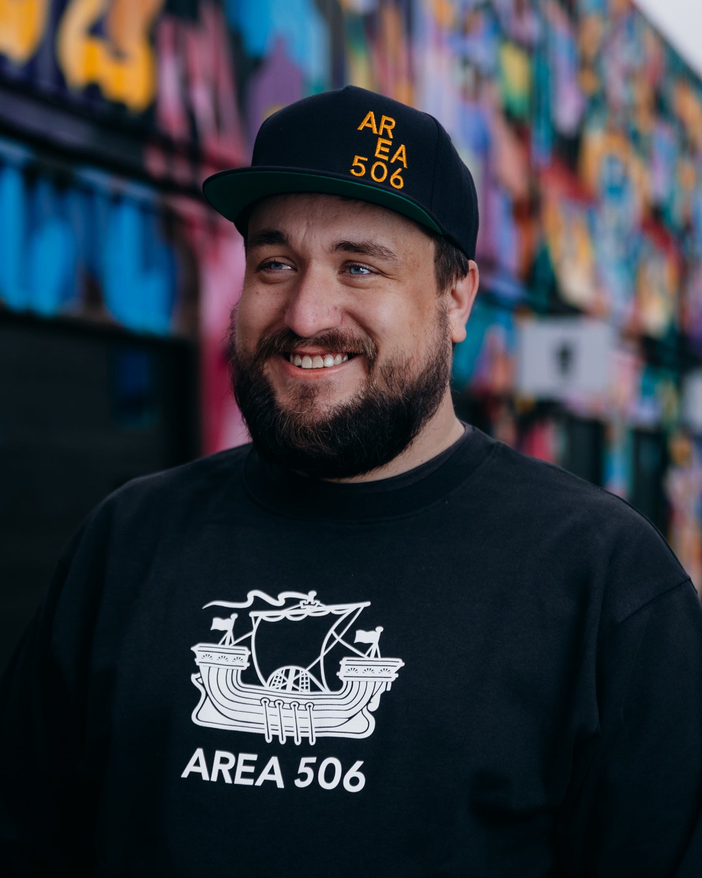 Man wearing a black 'AREA 506' cap and sweatshirt in front of a colourful mural.
