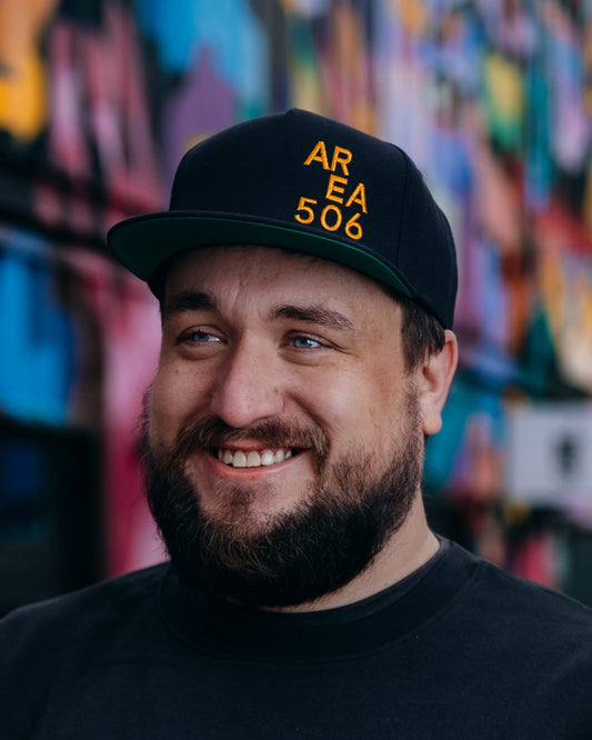 Man wearing a black 'AREA 506' cap and sweatshirt in front of a colourful mural.