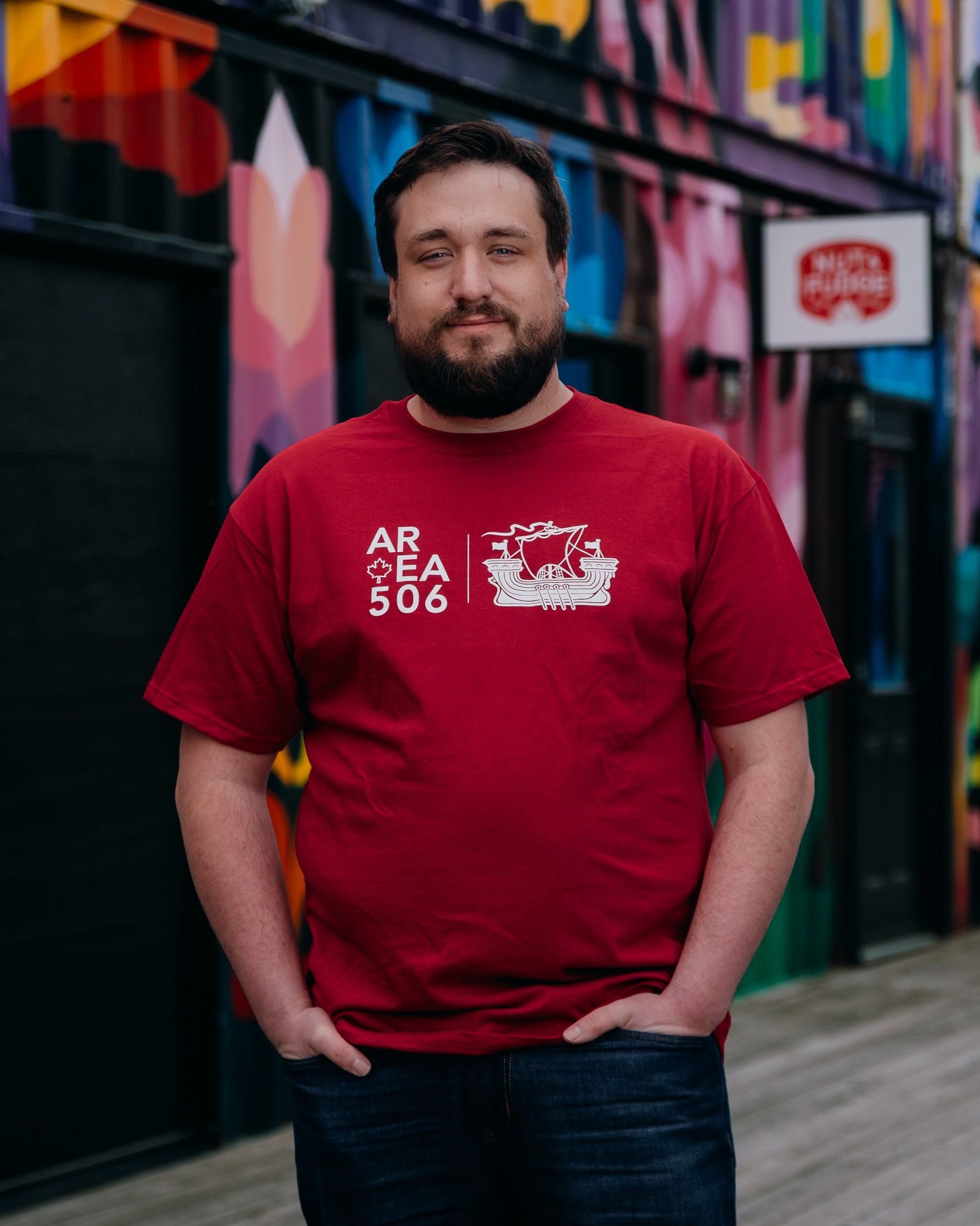 Man wearing a red t-shirt with a logo and text, standing in front of a colorful mural.
