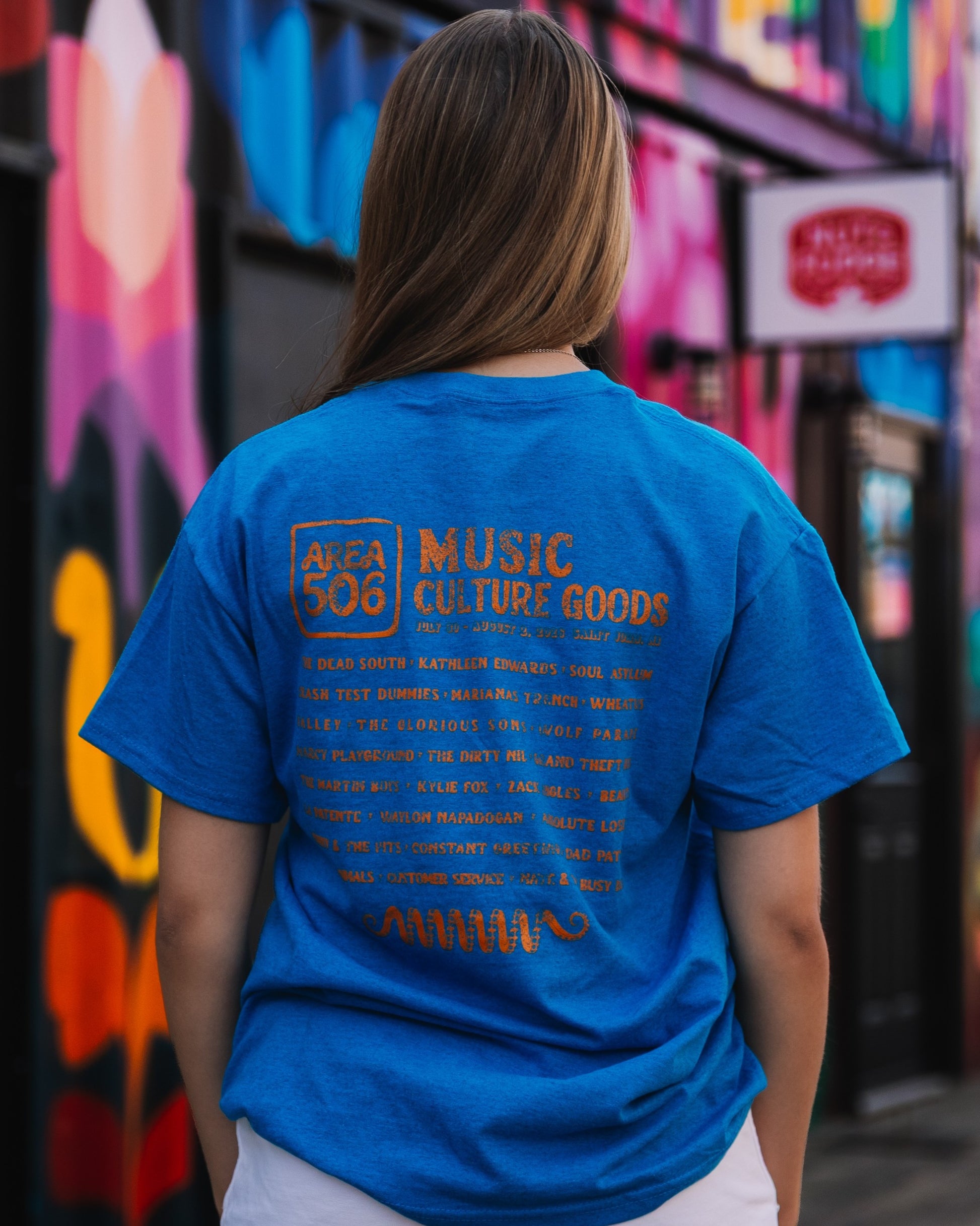 A woman viewed from behind wearing a bright blue graphic t-shirt with orange text that reads 'AREA 505 MUSIC CULTURE GOODS'. The background is a colourful, graffiti-covered street wall.