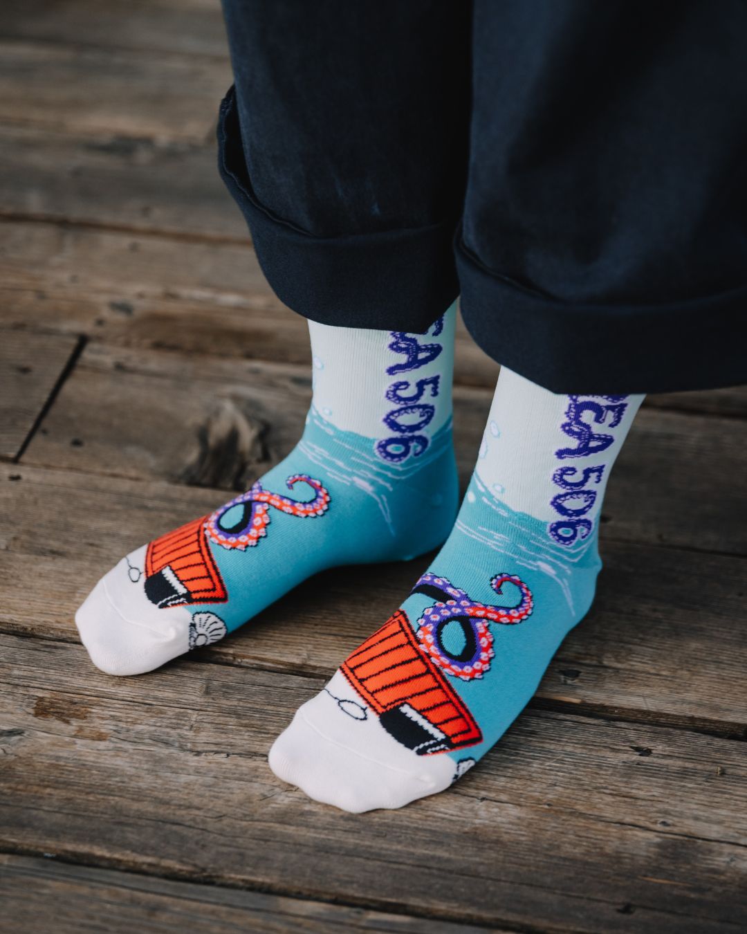 A person wearing white crew socks with a light blue lower section, purple vertical text reading 'AREA 506', and an orange-and-purple octopus tentacle graphic, standing on a rustic wooden plank surface.