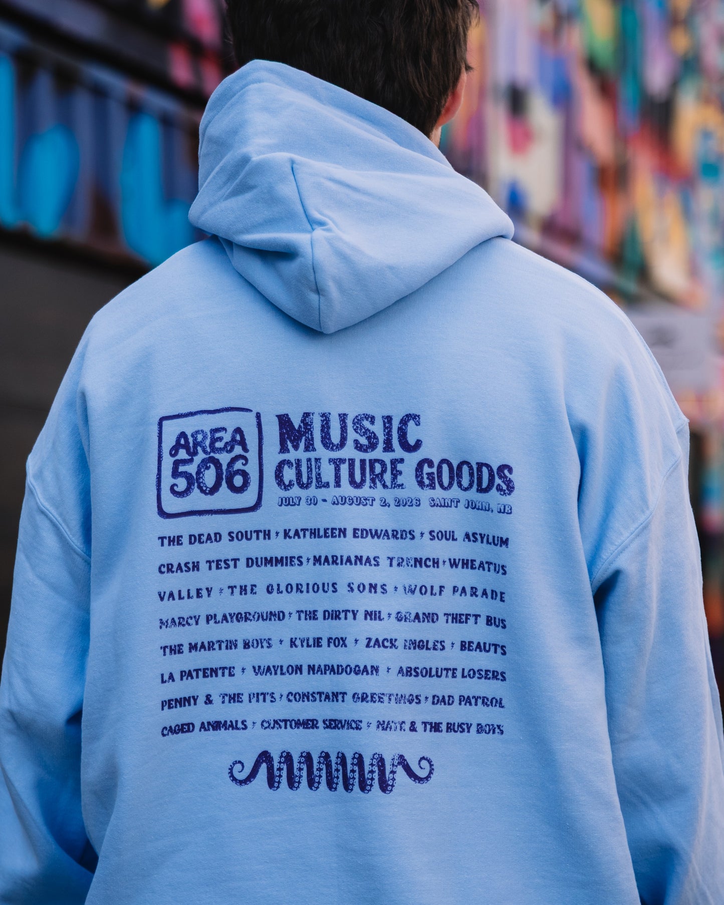 A person viewed from behind wearing a light blue hoodie with a large purple graphic print on the back. The print features the text 'AREA 506 MUSIC CULTURE GOODS' and a list of bands.