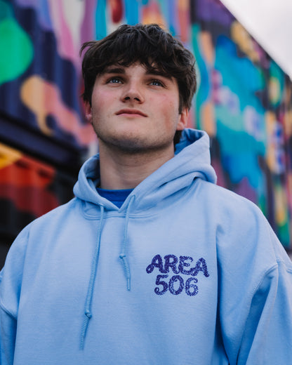 A young man wearing a light blue hoodie with 'AREA 506' printed in dark purple on the chest, standing in front of a colourful graffiti wall.