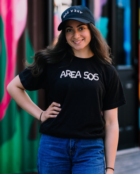 Woman wearing a black 'AREA 506' t-shirt and cap in front of a colourful mural.