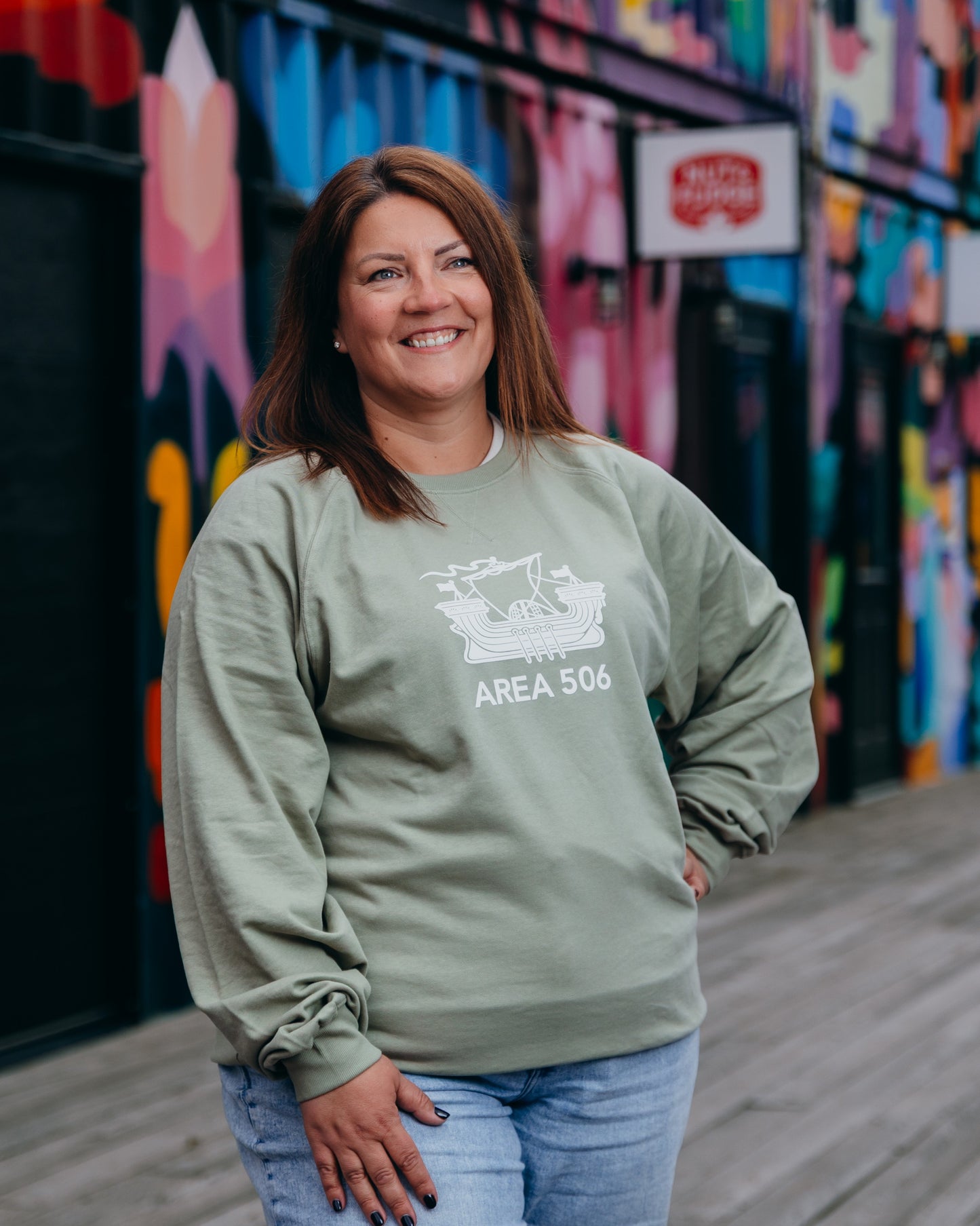 Woman wearing a green sweatshirt with 'AREA 506' text, standing in front of a colourful mural.