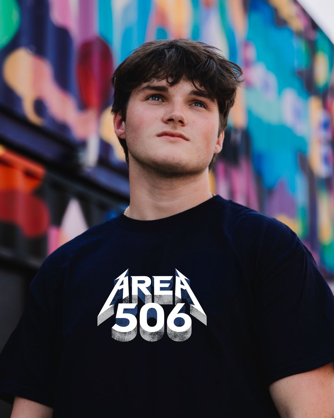 A young man wearing a dark navy t-shirt with a white 'AREA 506' graphic, standing in front of a colourful graffiti wall.