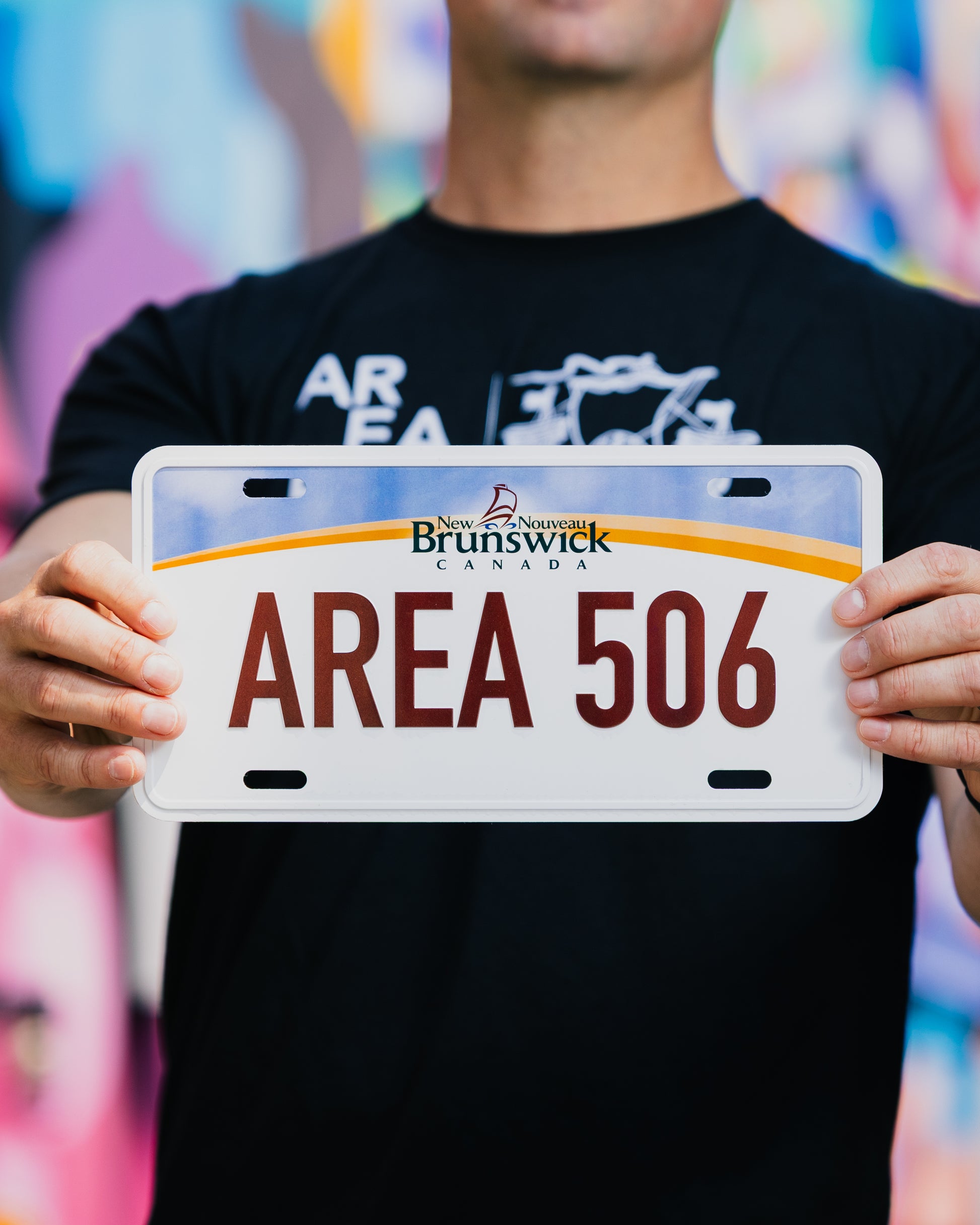 Person holding a tin sign shaped like a license plate with 'AREA 506' and New Brunswick branding.