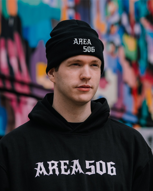 Person wearing a black 'AREA 506' beanie and hoodie in front of a colourful graffiti wall.