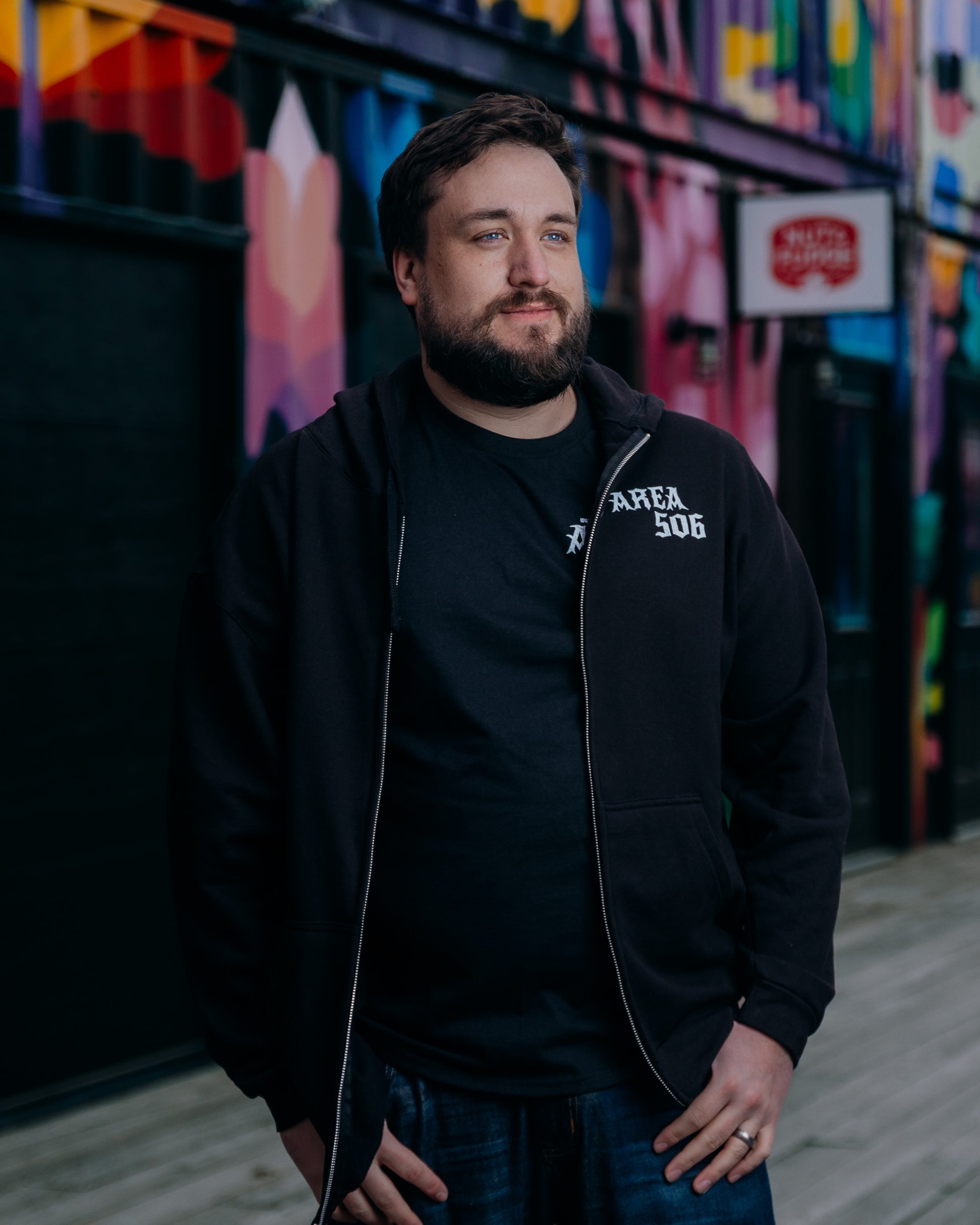 Man wearing a black hoodie with 'AREA 506' text in front of a colourful mural.