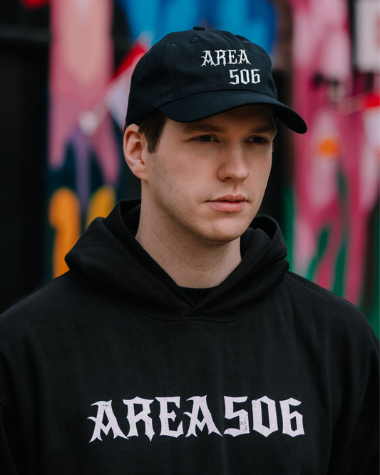 Person wearing a black 'AREA 506' cap and hoodie against a graffiti-covered wall.