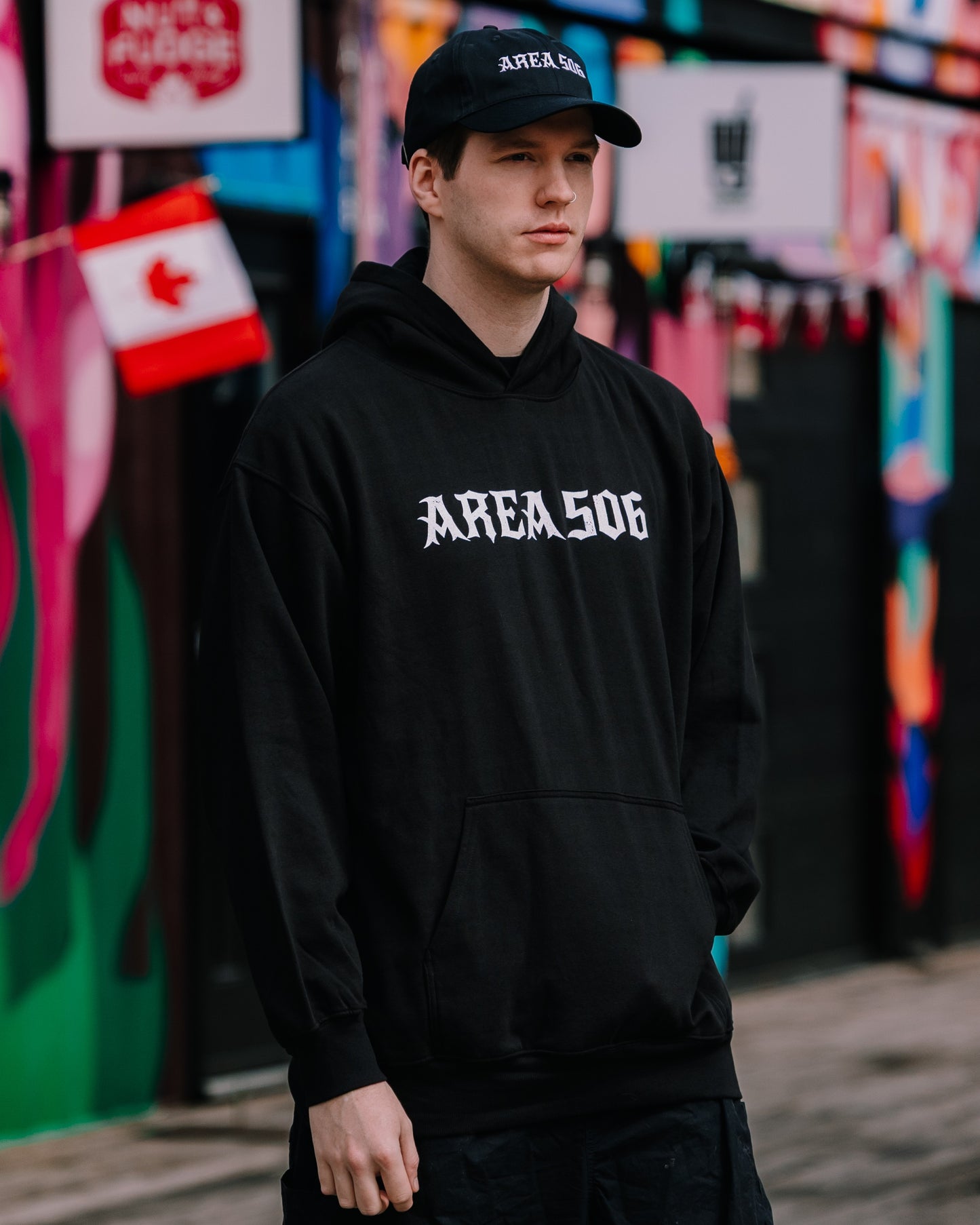 Person wearing a black hoodie with 'AREA506' white text in front of a colourful mural.