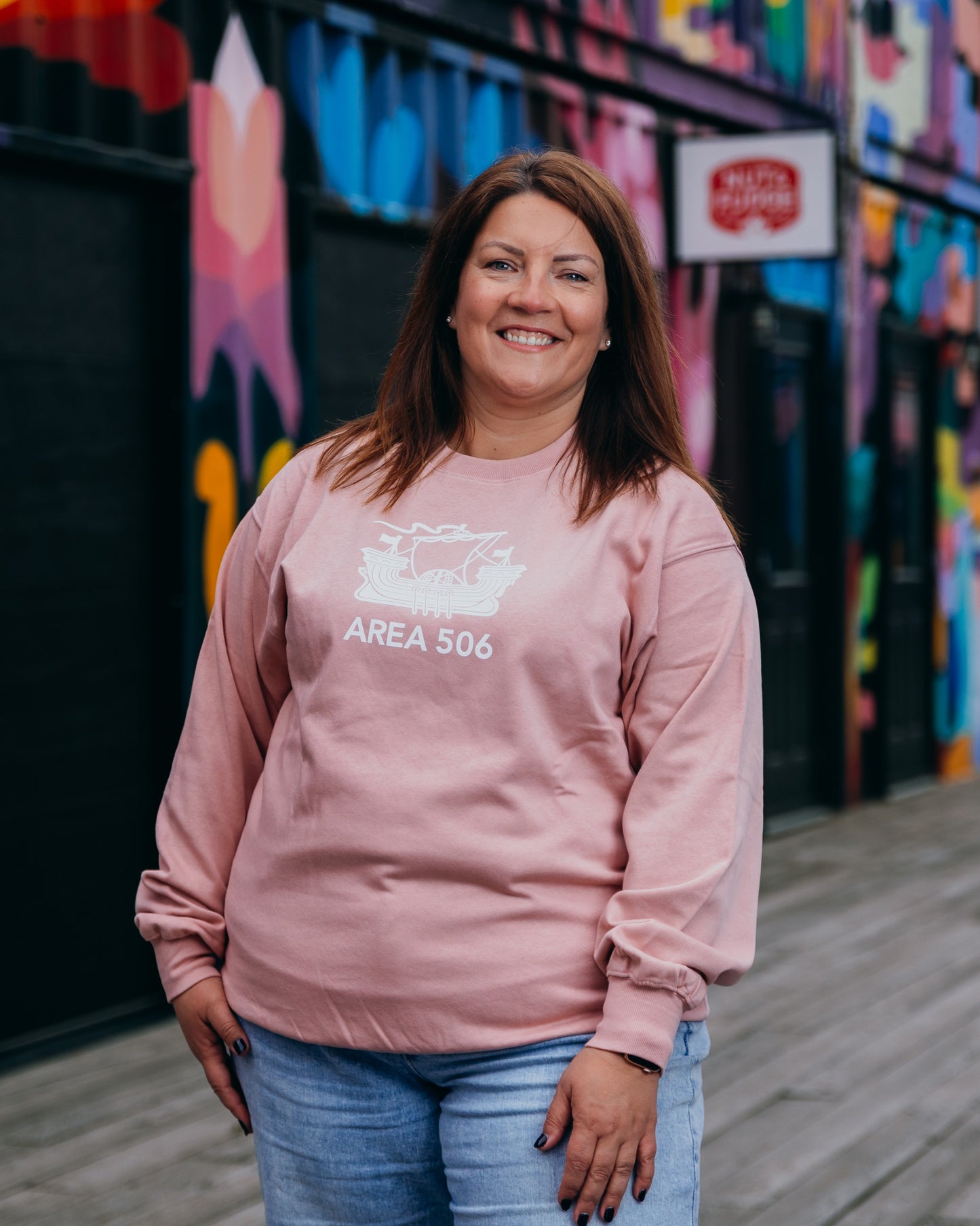 Woman wearing a pink sweatshirt with text, standing in front of a colourful mural.