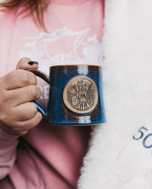 Navy mug with a bronze emblem held by a person wearing a pink sweater.