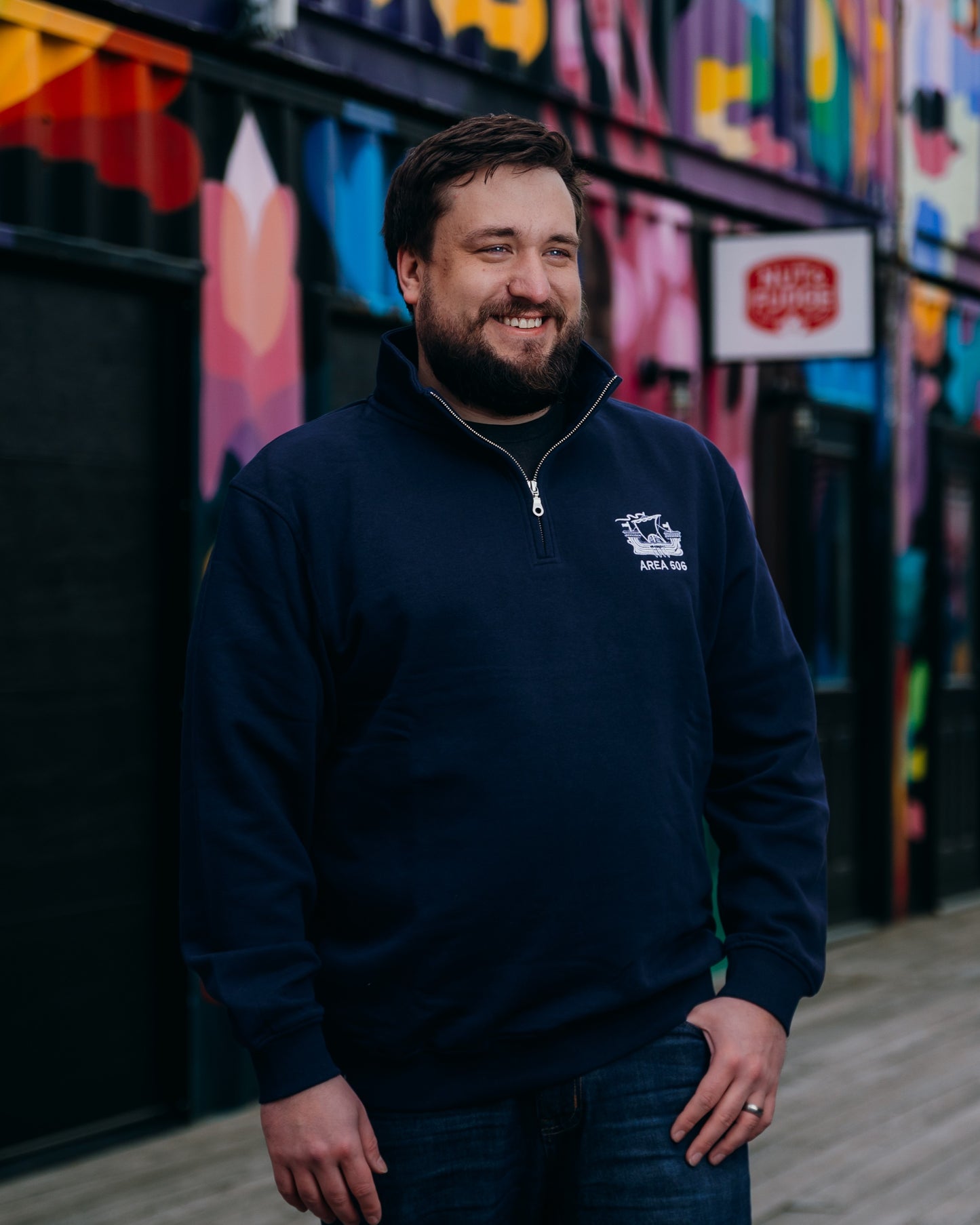 Man wearing a navy blue sweater with a logo standing in front of a colourful mural.