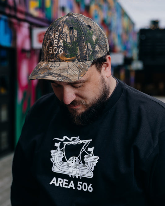 Person wearing a camouflage 'AREA 506' cap and black sweatshirt with logo