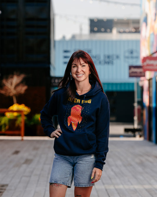 A person wearing a navy hoodie with a red and yellow rocket ship logo, standing in a public space with buildings in the background.