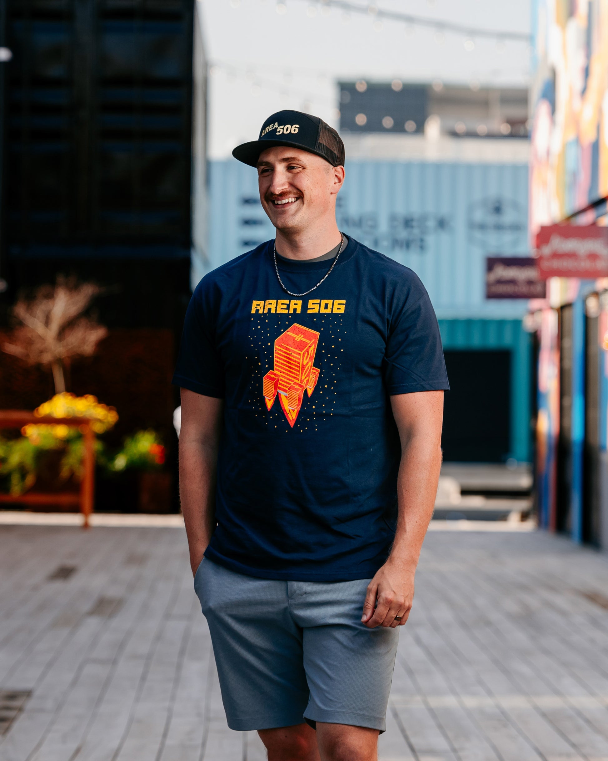 A person wearing a navy blue t-shirt with a red and yellow rocket ship logo, paired with light gray shorts and a black cap, standing on a street with buildings in the background.