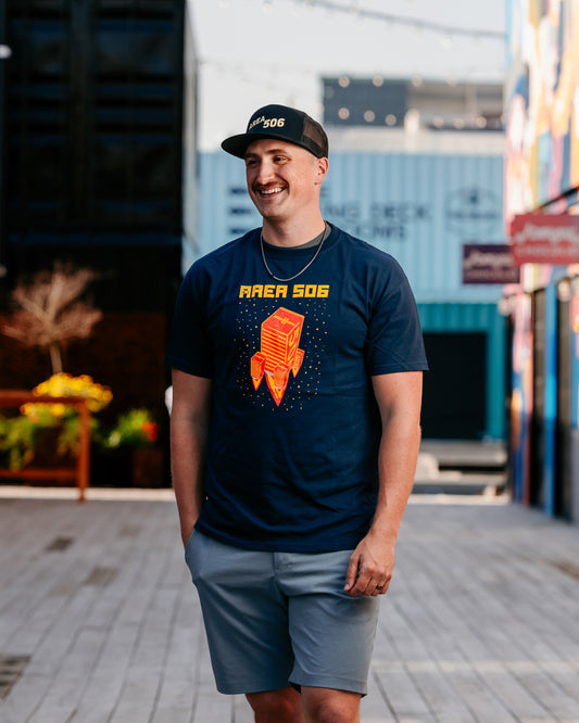 A person wearing a navy blue t-shirt with a red and yellow rocket ship logo, paired with light gray shorts and a black cap, standing on a street with buildings in the background.