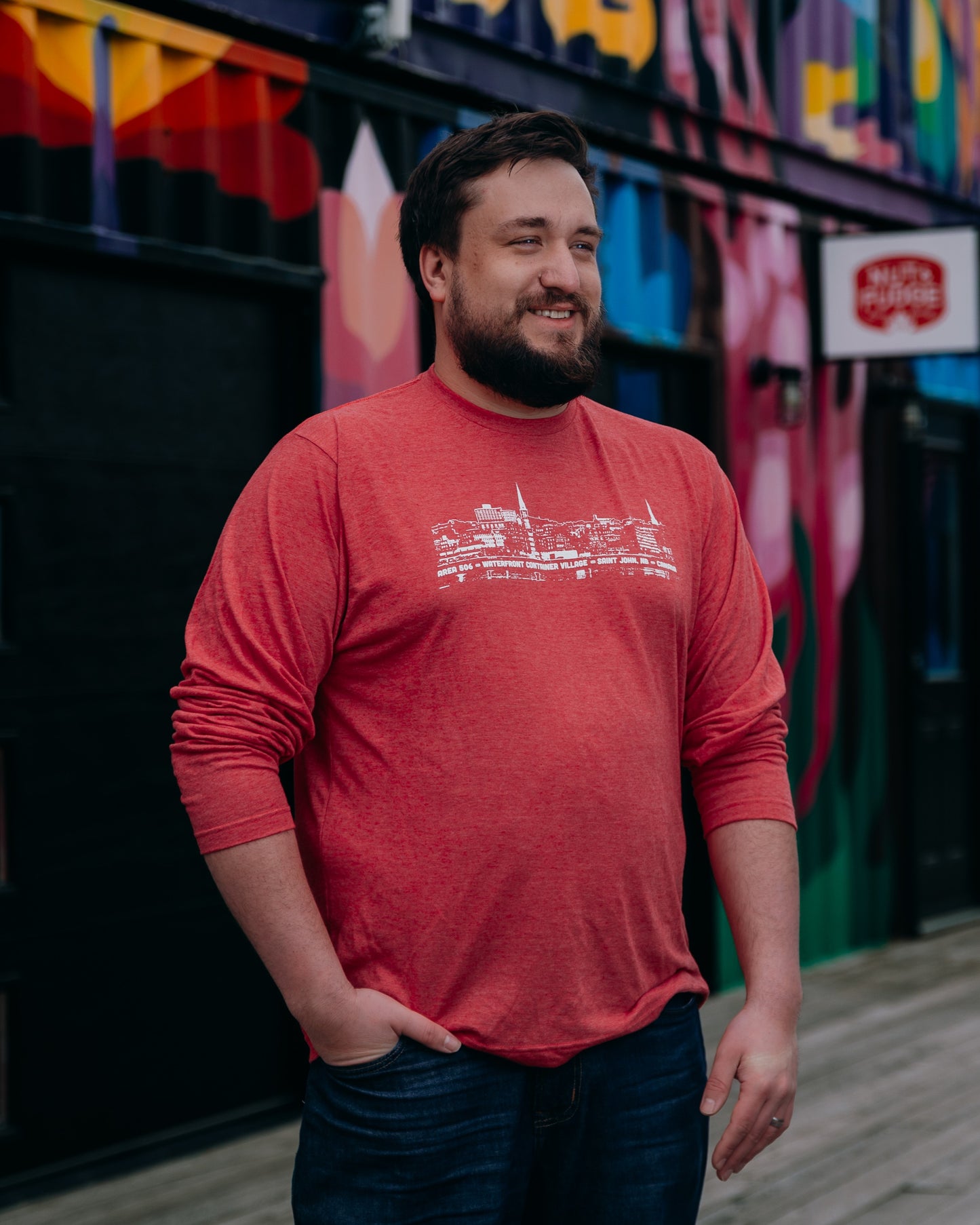 Man wearing a red shirt with white line drawing, standing in front of a colourful mural.