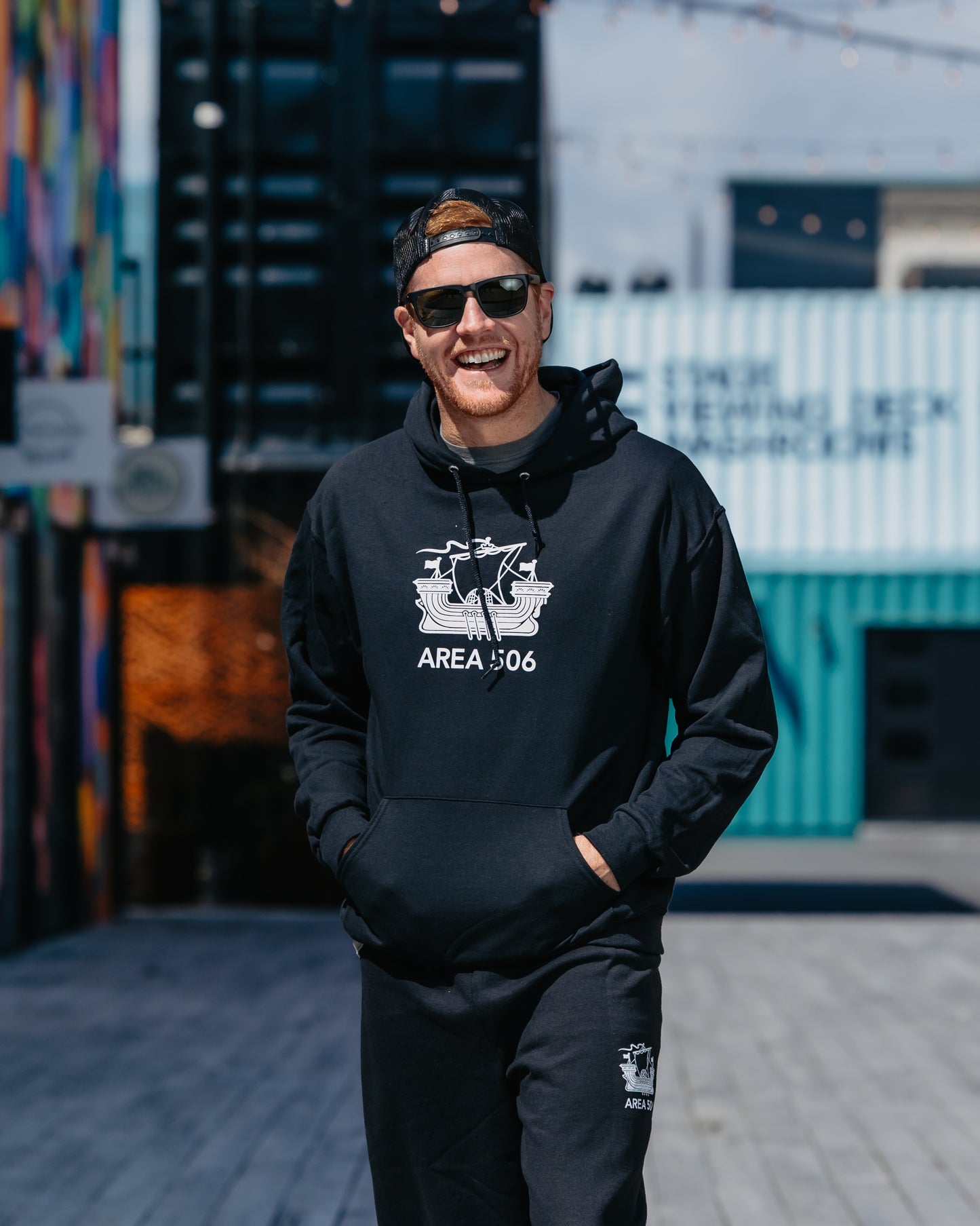 A person wearing a blue pullover hoodie with a white logo that reads 'AREA 506', standing in front of a colourful wall with graffiti.