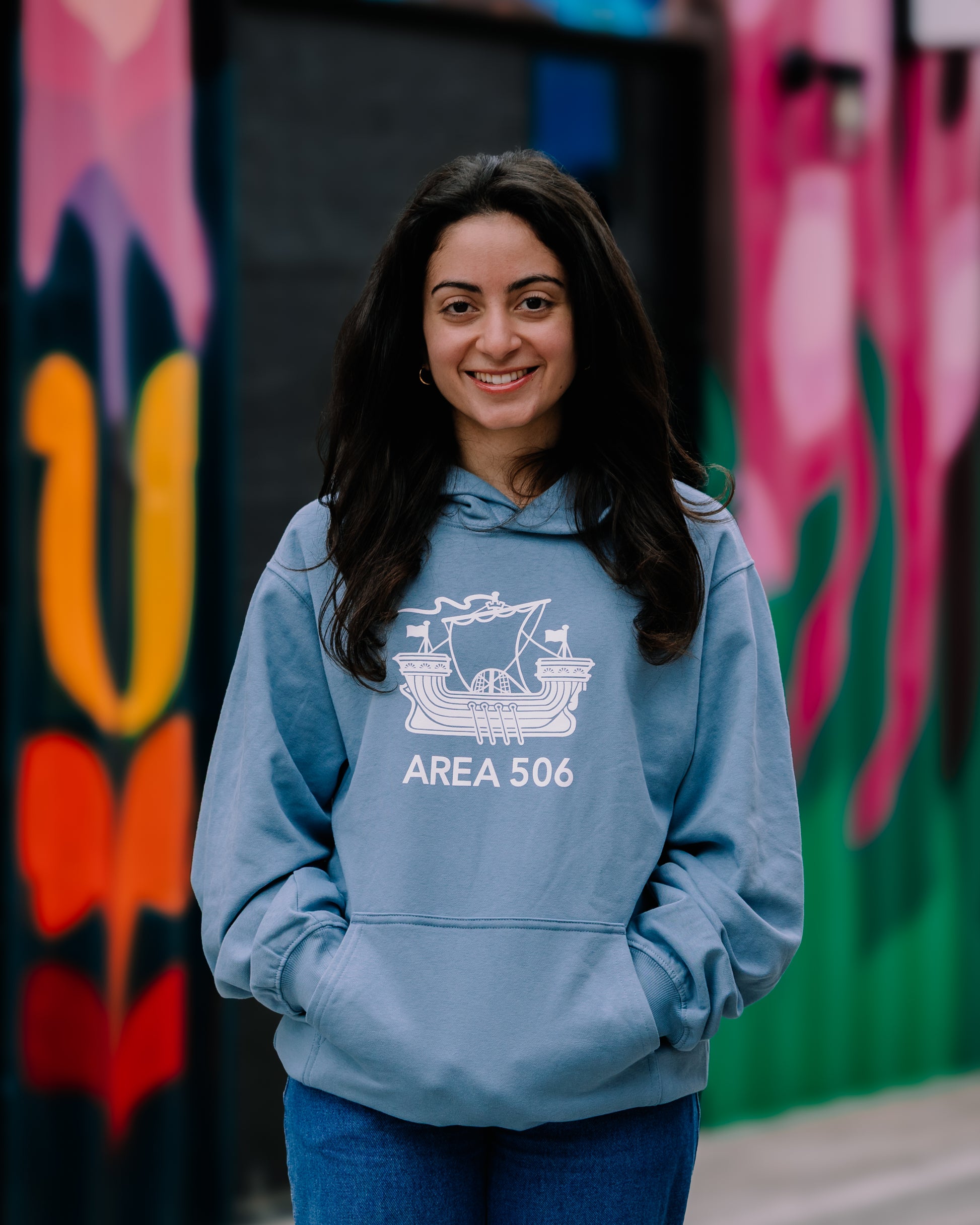 A person wearing a blue pullover hoodie with a white logo that reads 'AREA 506', standing in front of a colourful wall with graffiti.