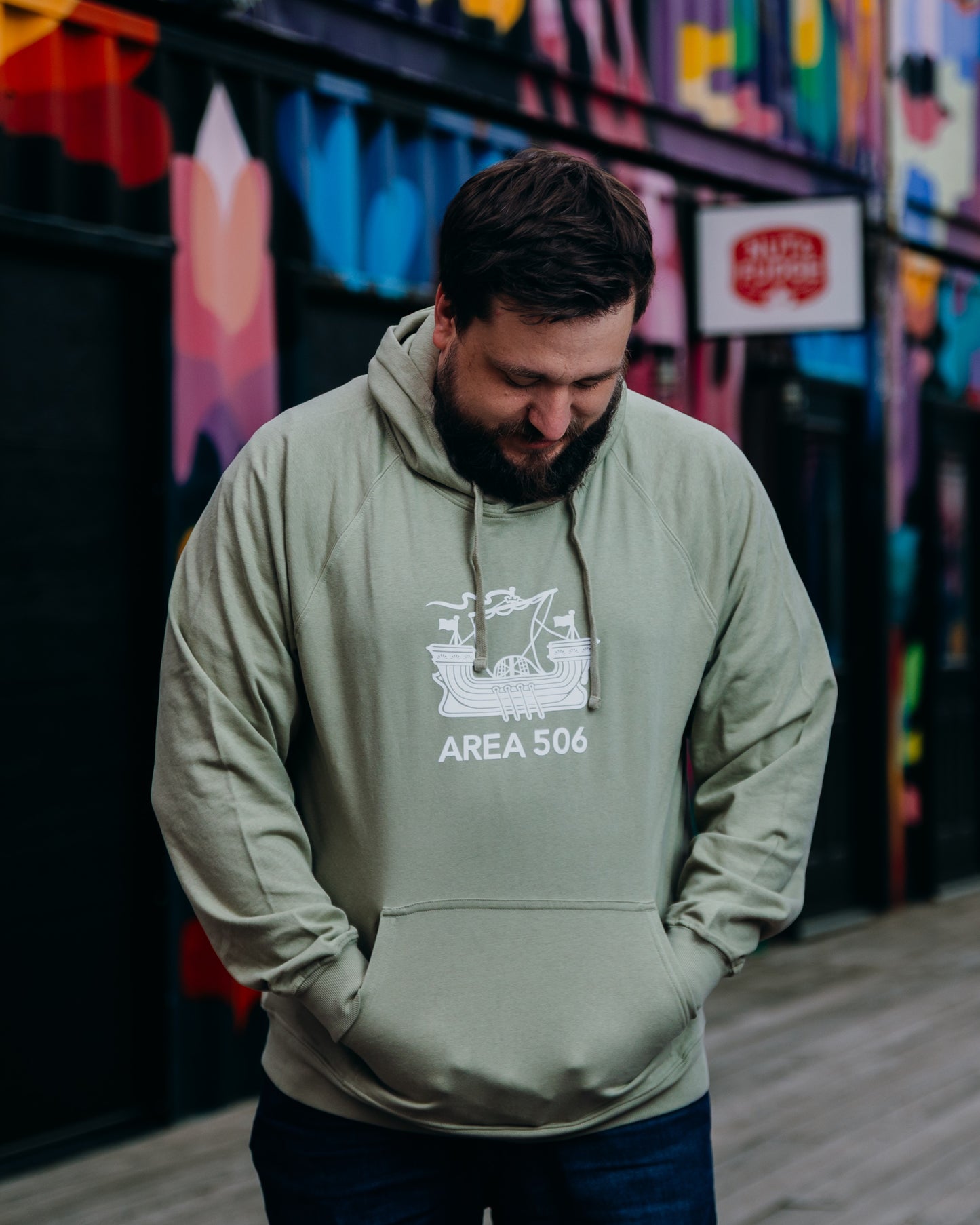 Man wearing a green hoodie with 'AREA 506' logo in front of a colourful mural.