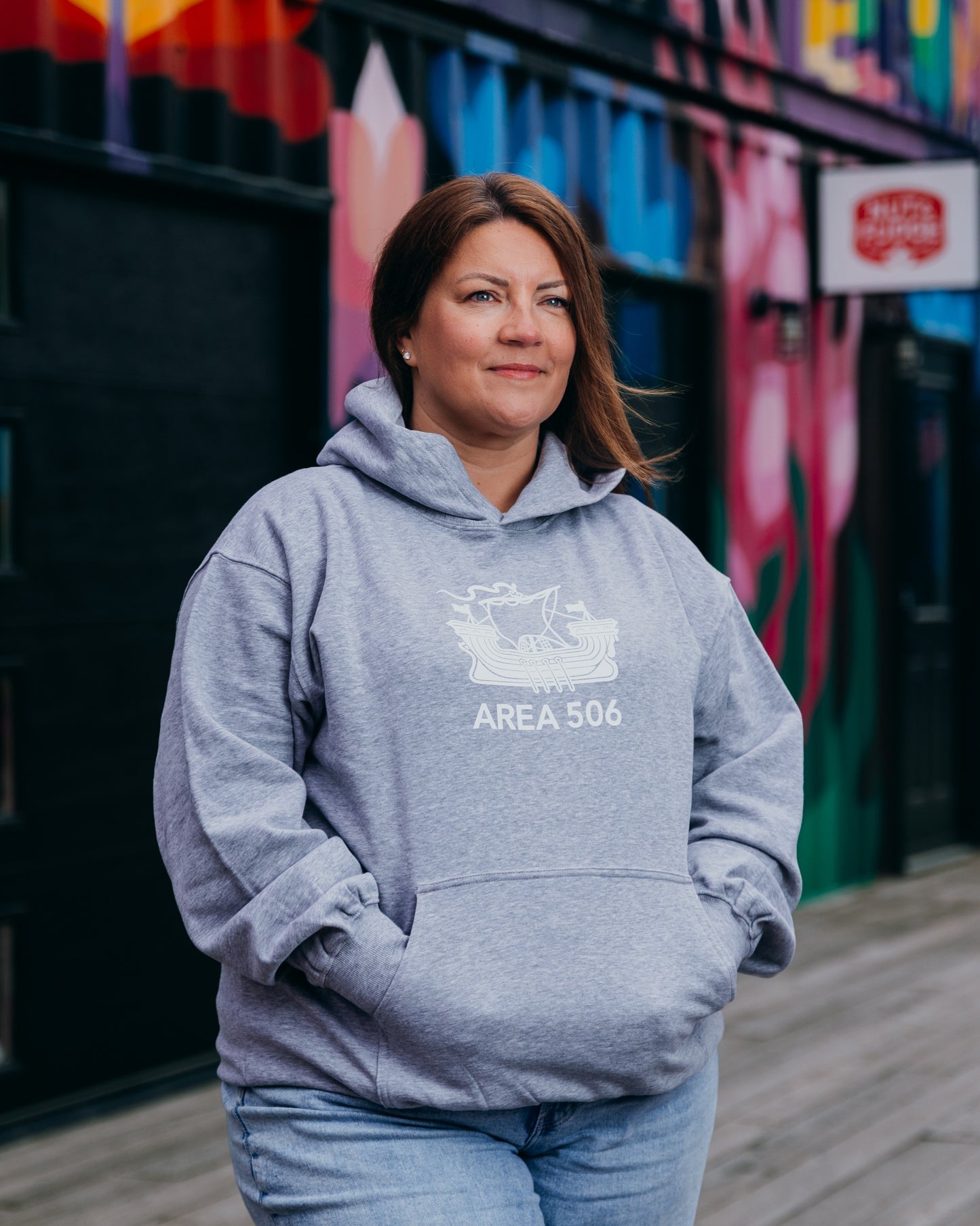 Woman wearing a grey hoodie with 'AREA 506' design in front of a colourful mural.
