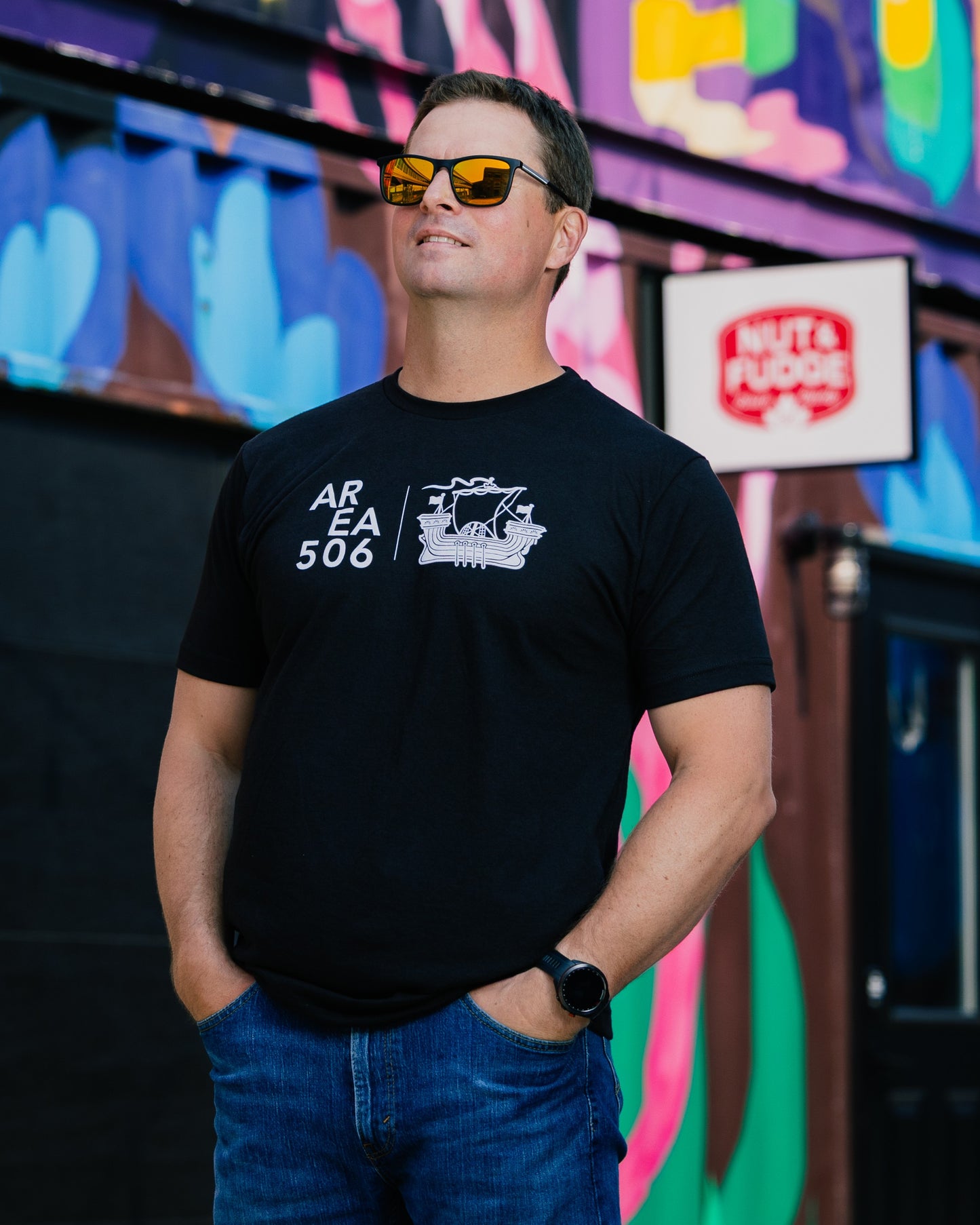 Man wearing a black t-shirt with white text and graphics, standing in front of colourful signs.
