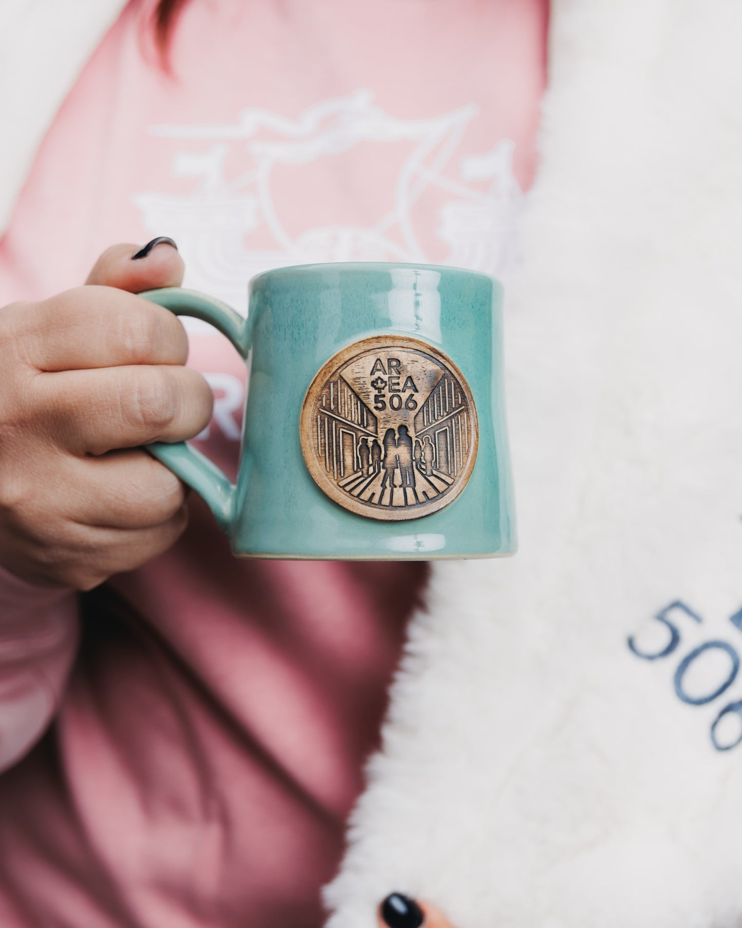 Person holding a green mug with a logo, wearing a pink shirt and white blanket.