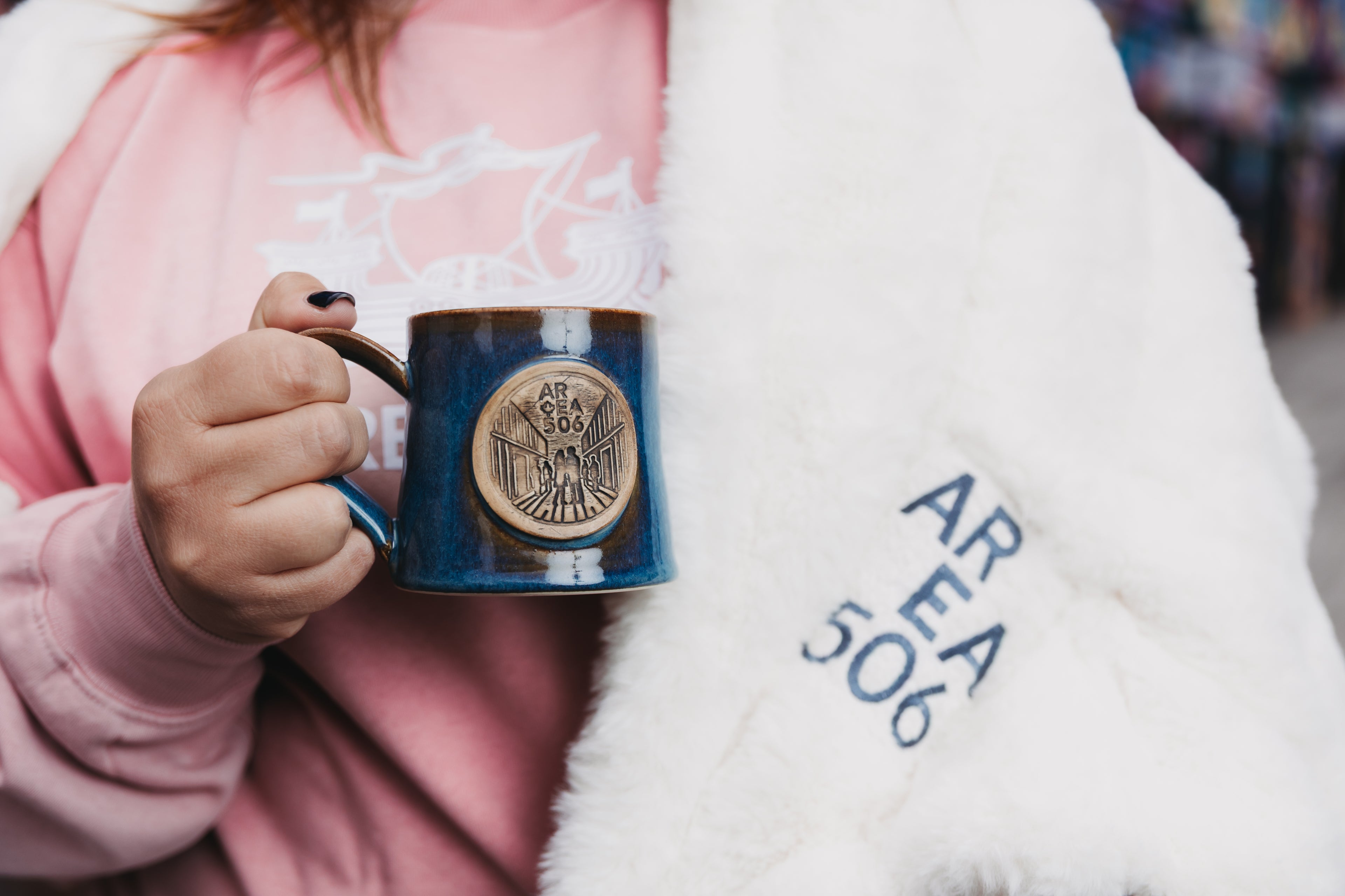 Person holding a blue mug with 'AREA 506' branding, wearing a pink shirt and white blanket.