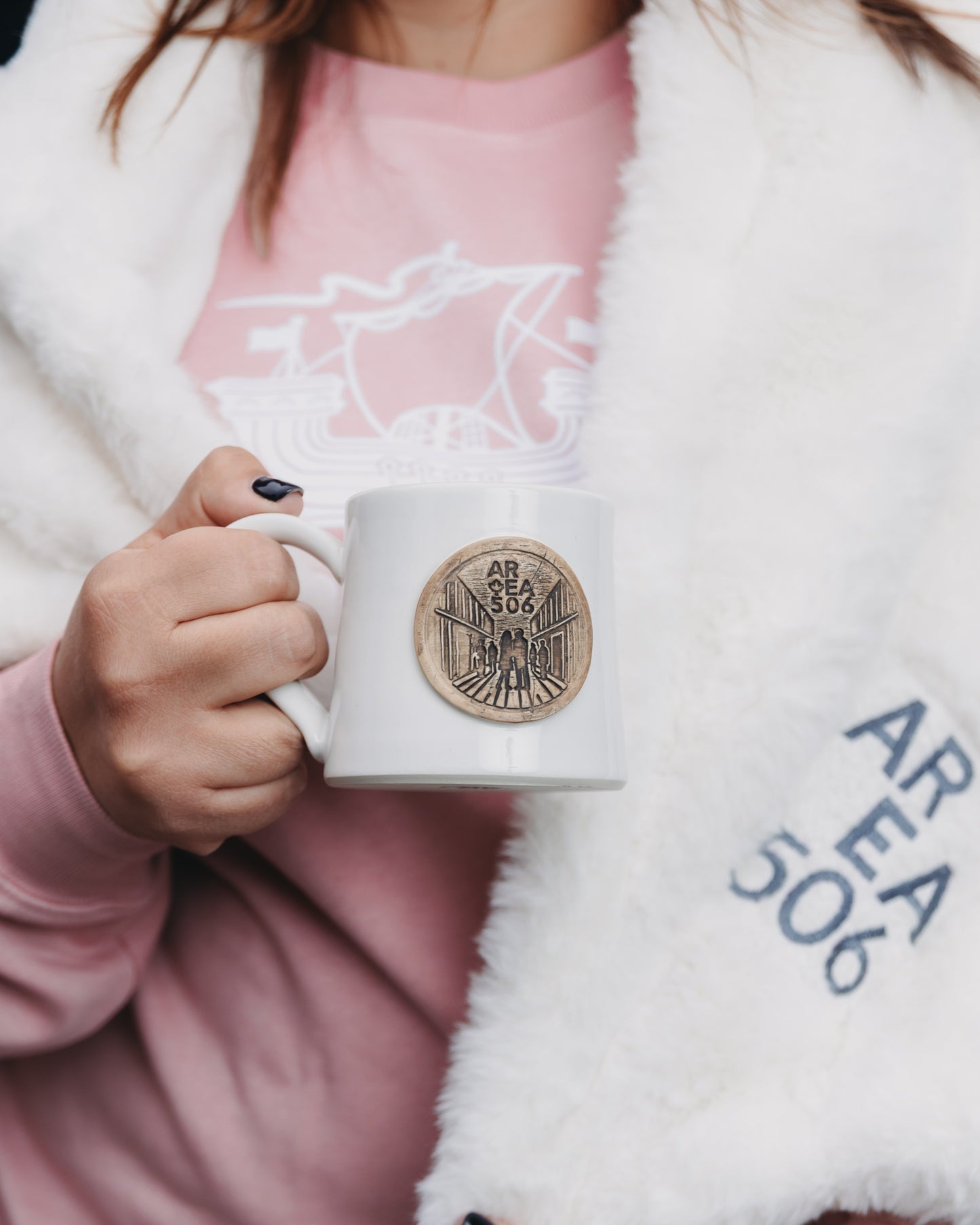 Person holding a white mug with a logo, wearing a pink shirt and white blanket.