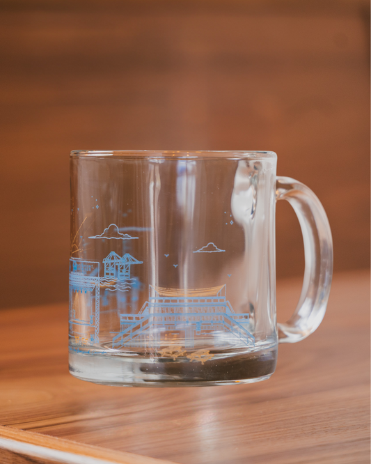 Clear glass mug with blue etched design of the Waterfront Container Village on a wooden surface.