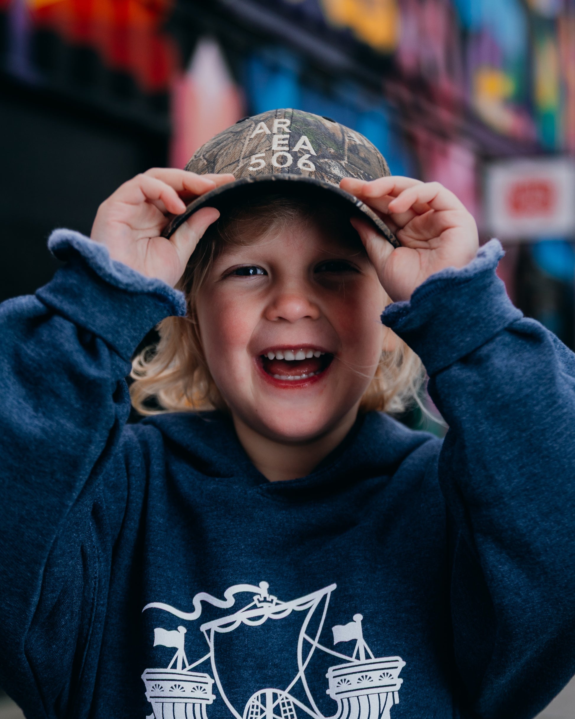 Child wearing a cap with 'AREA 506' text, smiling in front of a colourful mural.